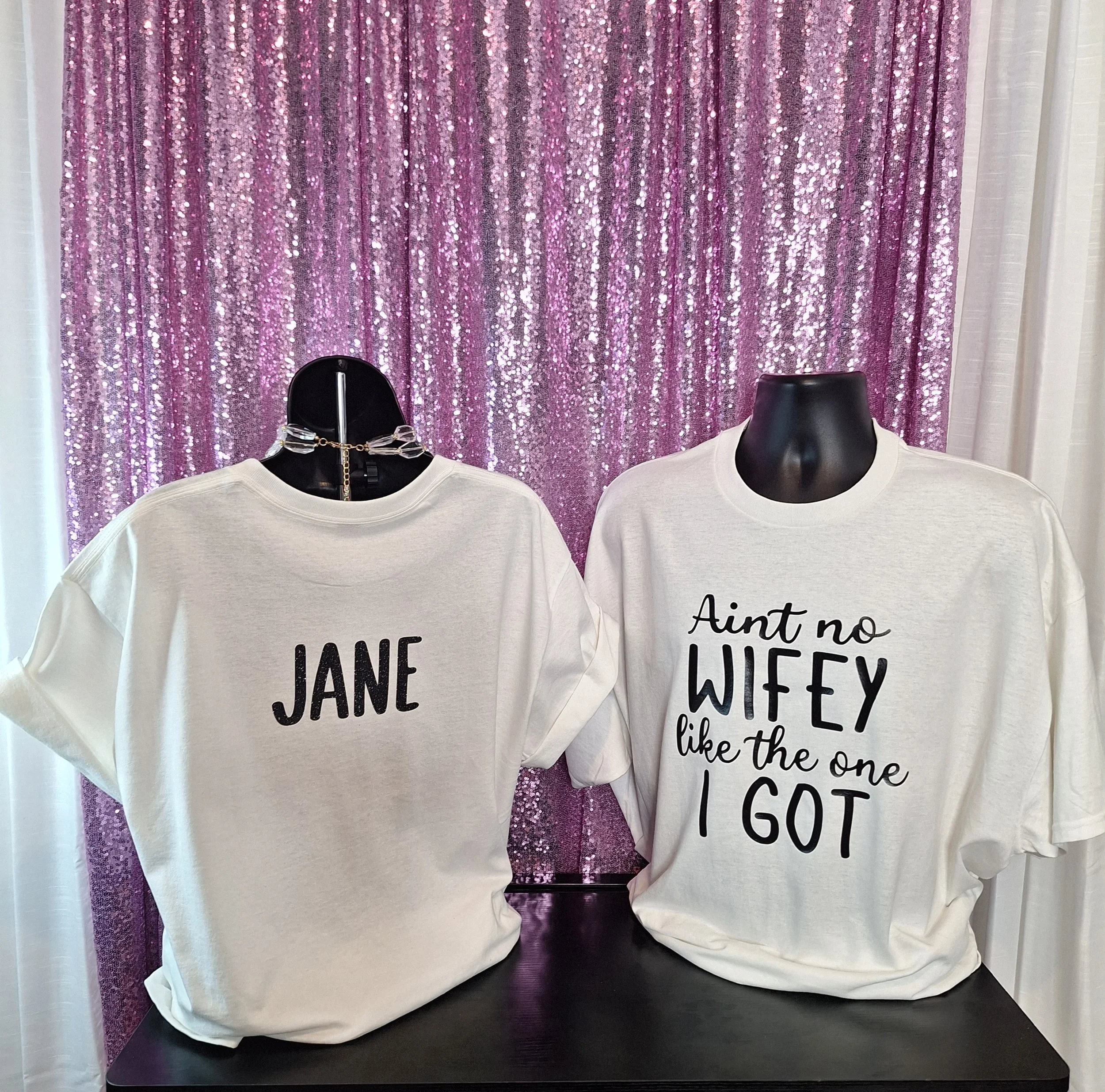 Mannequins wearing white T-shirts with black text, placed in front of a purple sequin backdrop.