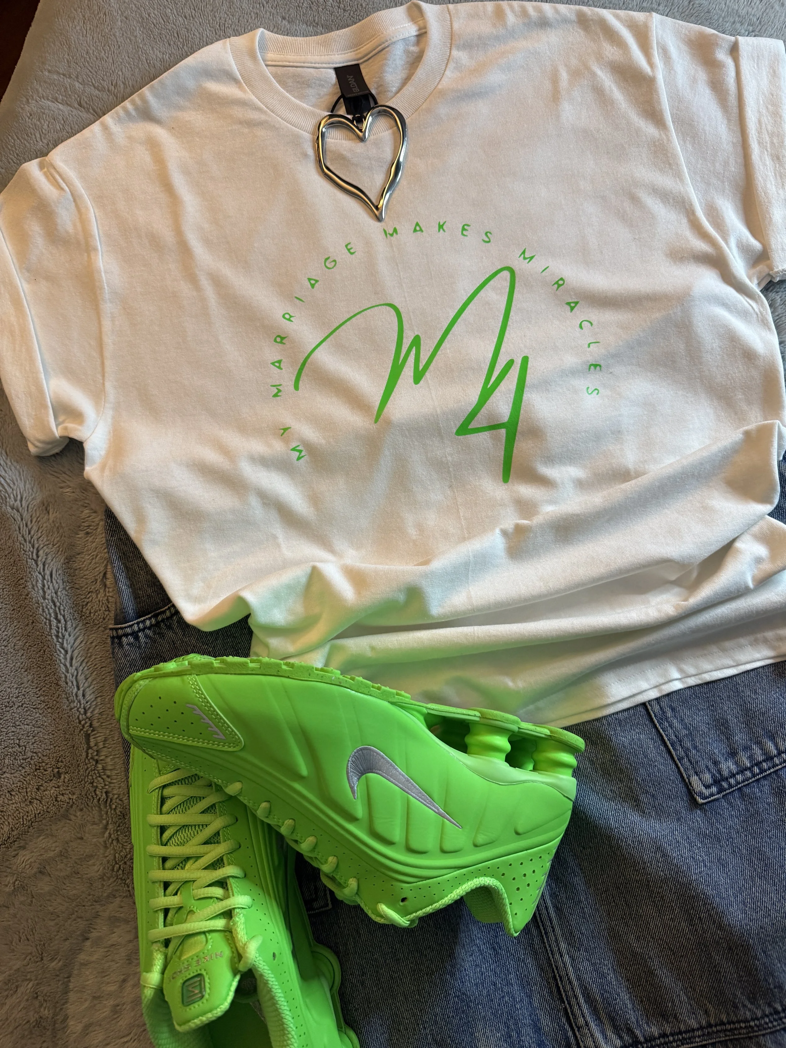 A white t-shirt with a green Nike sneaker and shorts. The t-shirt has a green circle with the words "WHAT MARRIAGE MAKE MIRACLE" and a large green signature-like letter 'M' in the center. The shirt is accessorized with a silver heart-shaped necklace.