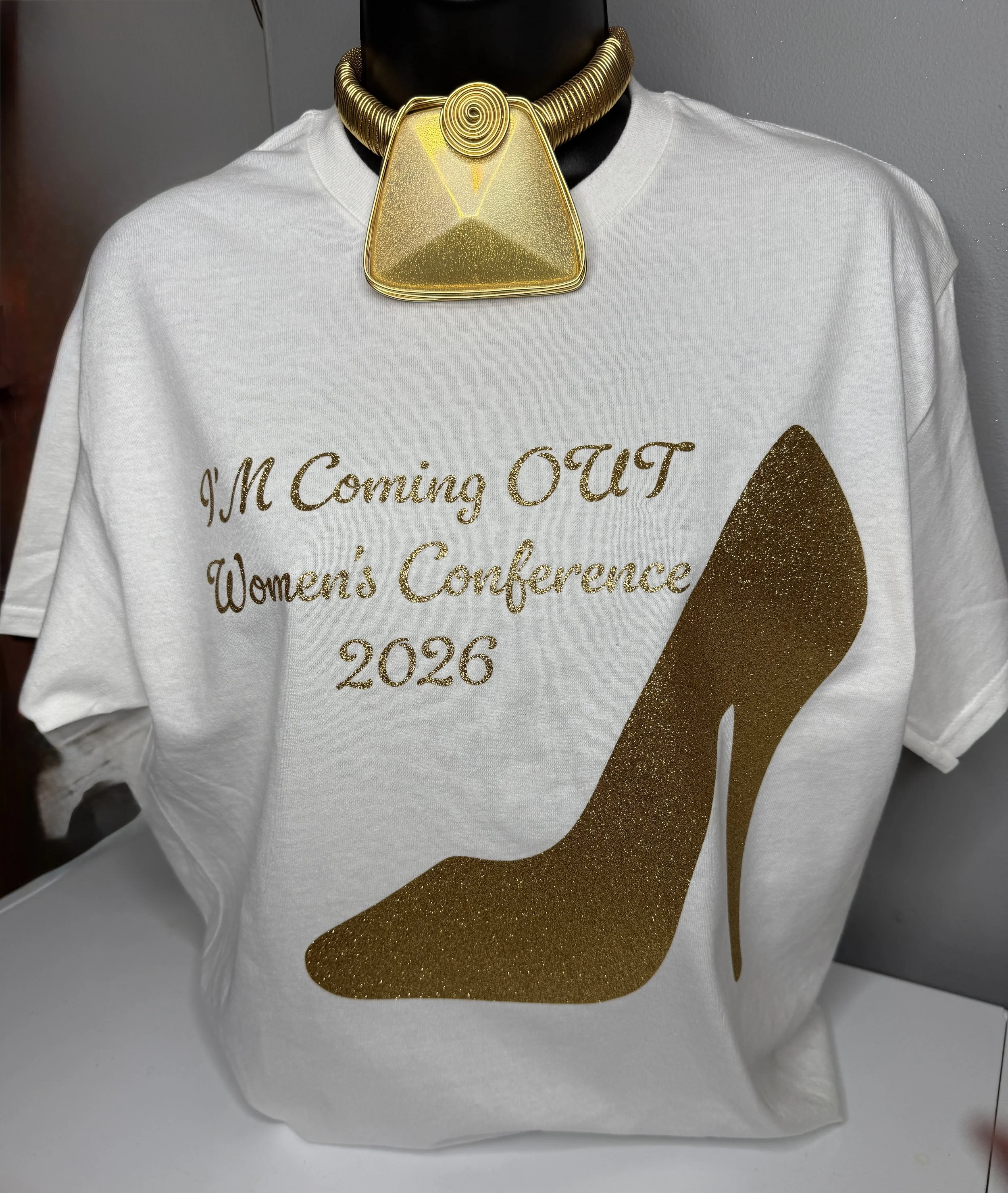 Gold statement necklace with a large triangular pendant, worn over a white T-shirt with gold glittery text reading 'I'M Coming OUT Women's Conference 2026'.
