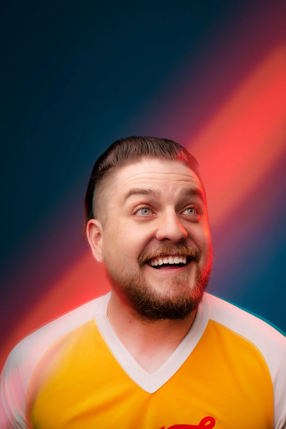 A young man with a beard and mustache smiling and looking upward. He is wearing a yellow and white shirt against a dark background with a red and blue light gradient.