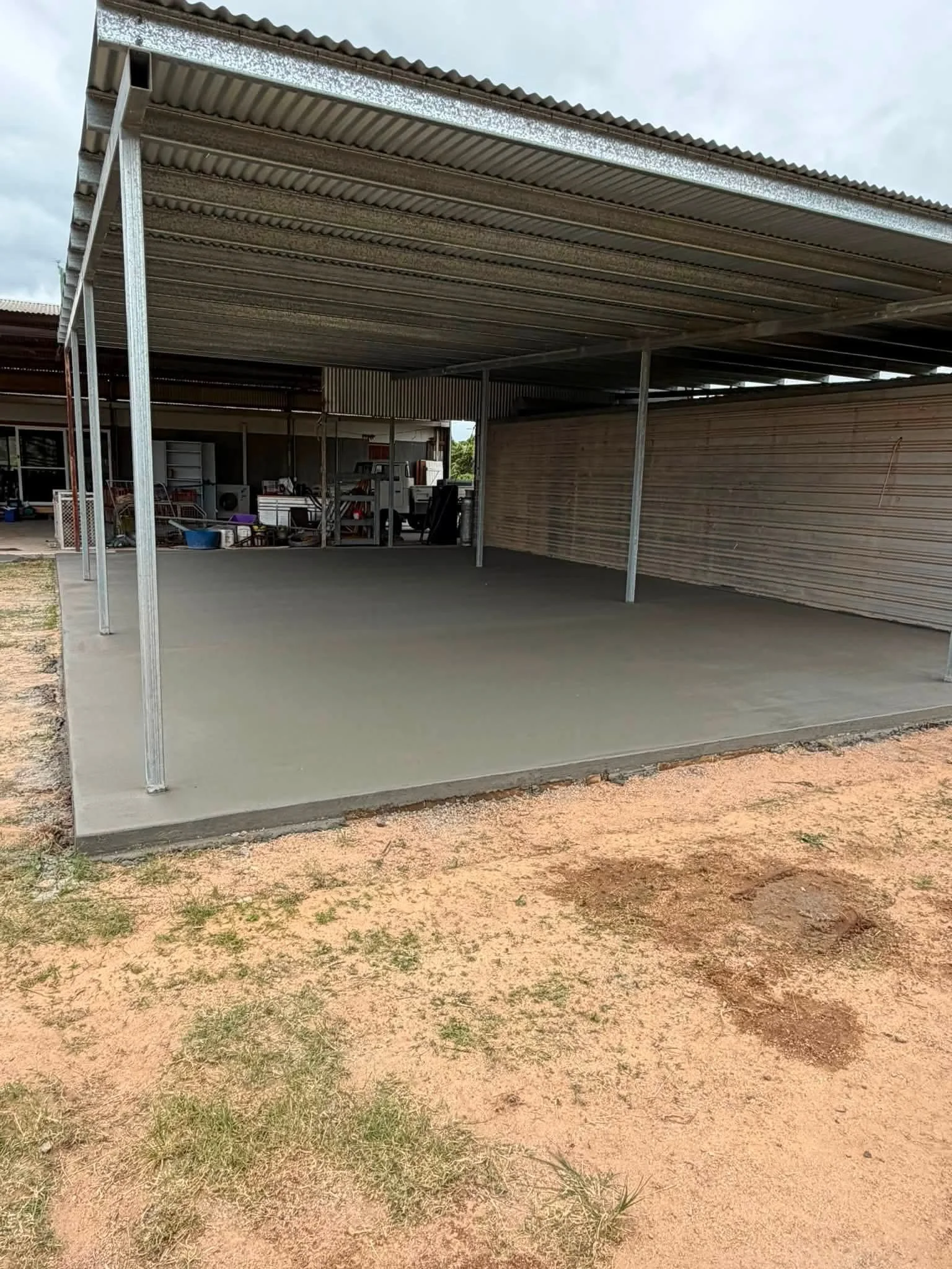 Empty construction site with a concrete slab floor and metal roofing supported by metal columns. There are various building materials and equipment in the background, and the ground is a mixture of dirt and patches of grass.