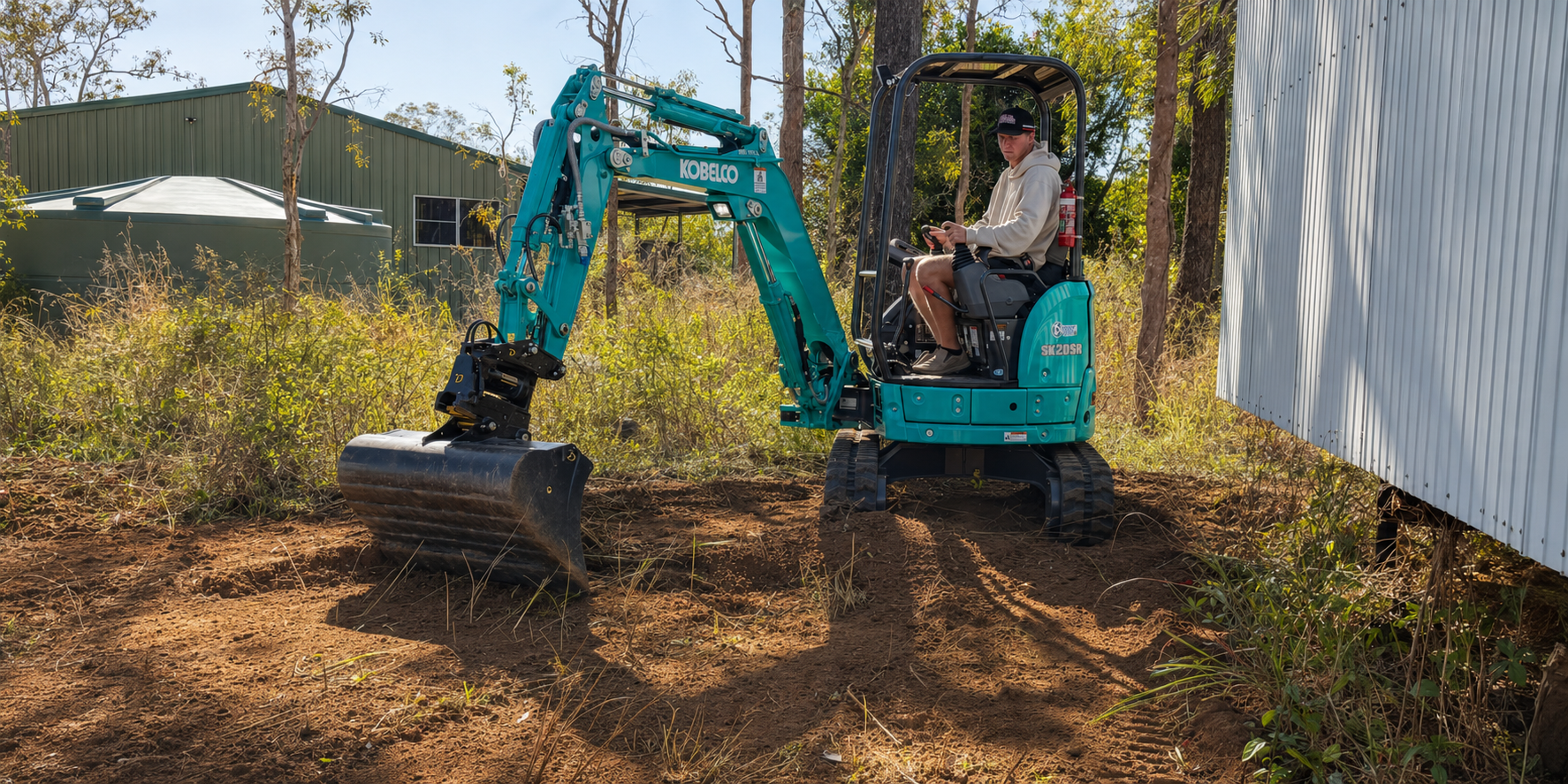 A person operating a blue mini excavator on a construction site with dirt and overgrown vegetation, near a green metal building and trees in the background.