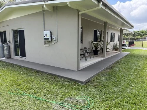 The back exterior of a house with a small covered porch, potted plants, and a grassy yard, with a green garden hose on the lawn.