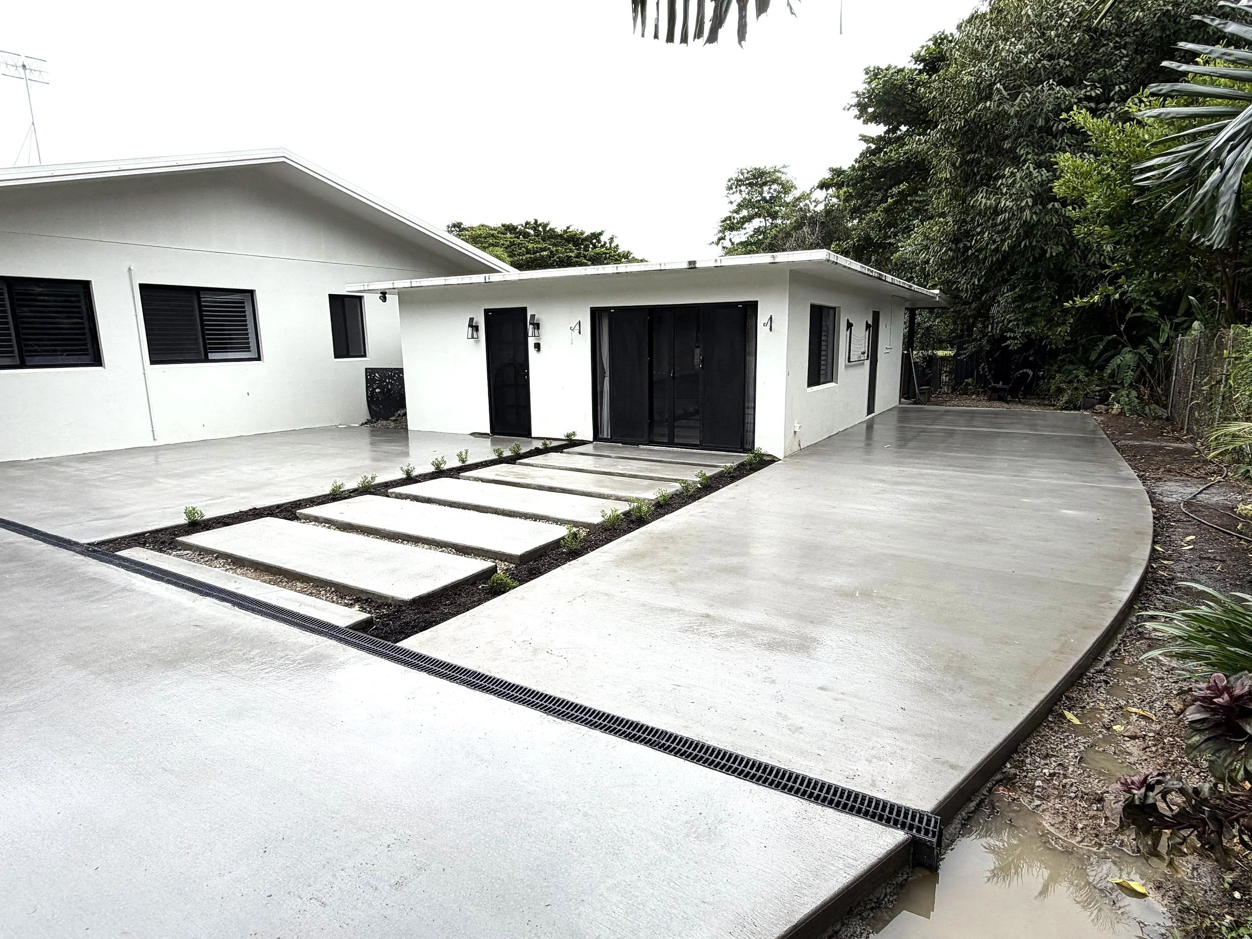 A modern house with a white exterior, black windows, and a flat roof. There is a concrete walkway leading to the house with small plants along the edge. The background has lush green trees and a cloudy sky.
