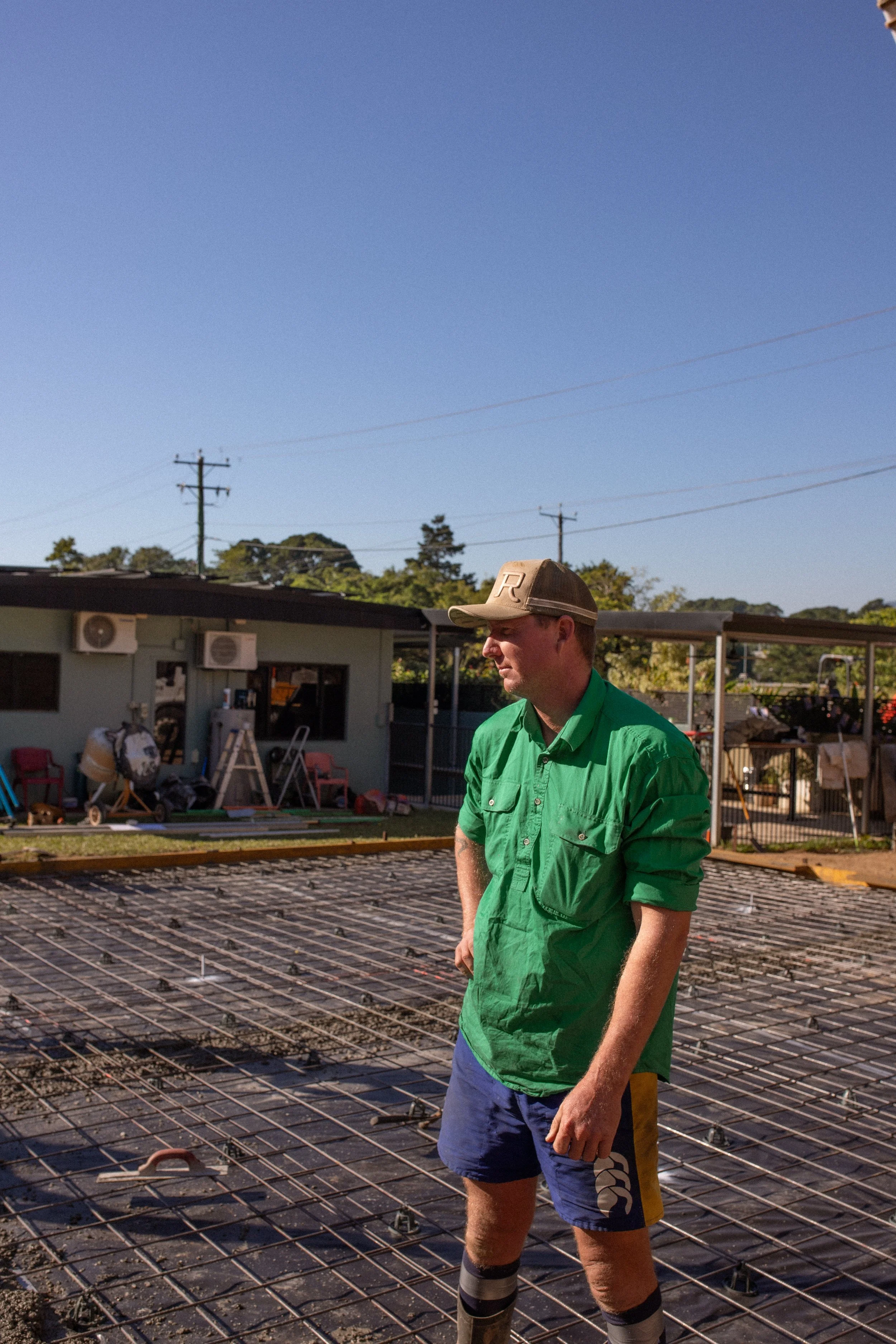 A man in a green shirt, blue shorts, and a beige cap standing on a construction site with metal rebar on the ground and residential buildings in the background.