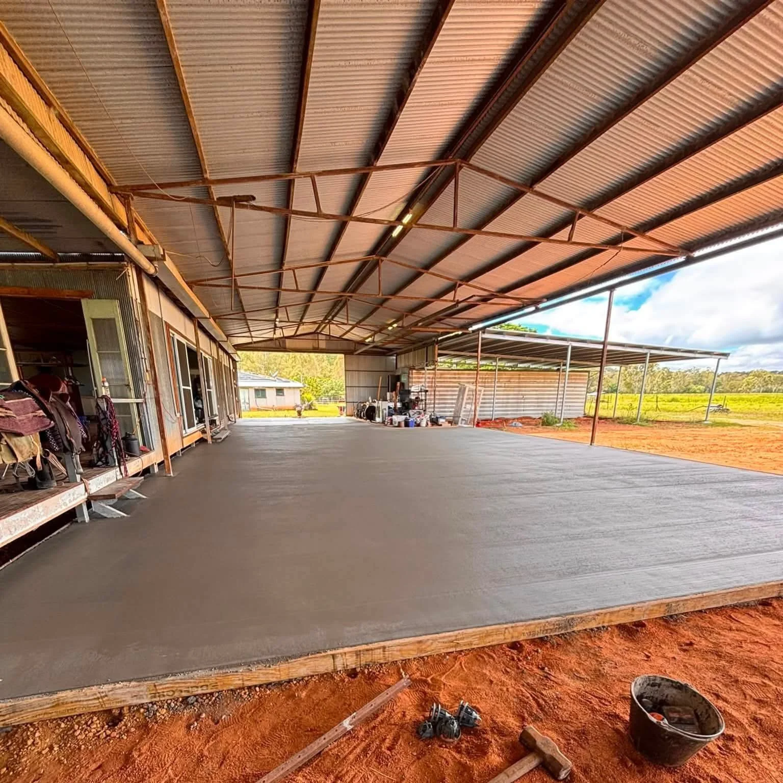 A partially constructed building with a concrete floor, metal roofing, and an open view to a field with grass and trees in the background. Construction tools and materials are scattered around.