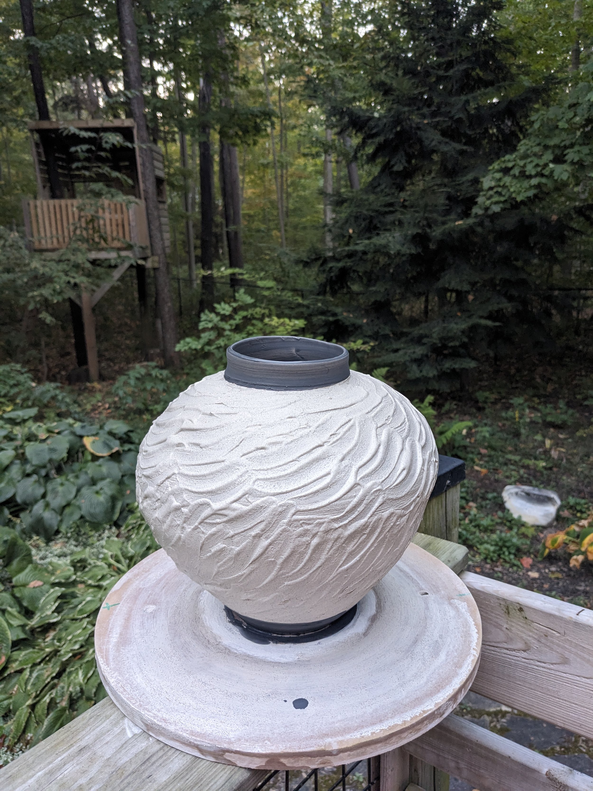 A handcrafted ceramic lamp with a textured surface, placed outdoors on a wooden table in a wooded area.