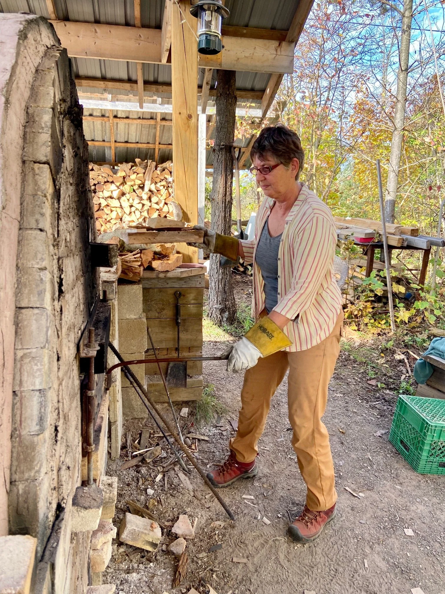 A woman working outdoors near a woodpile, wearing gloves, glasses, a striped shirt, tan pants, and red hiking shoes, in front of a small wood-fired oven or kiln, with trees and a utility table in the background.