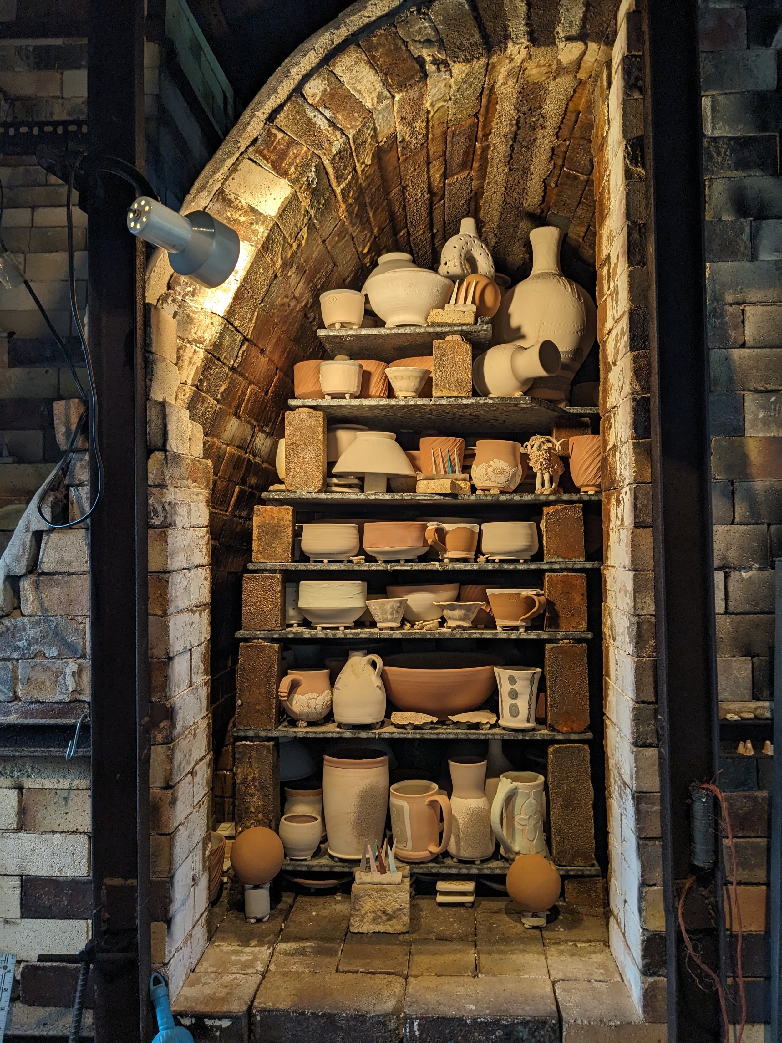 A display of various ceramic pottery such as vases, bowls, and jugs arranged on shelves within a brick fireplace or alcove. The setting appears to be a pottery studio or display area with warm lighting highlighting the ceramics.