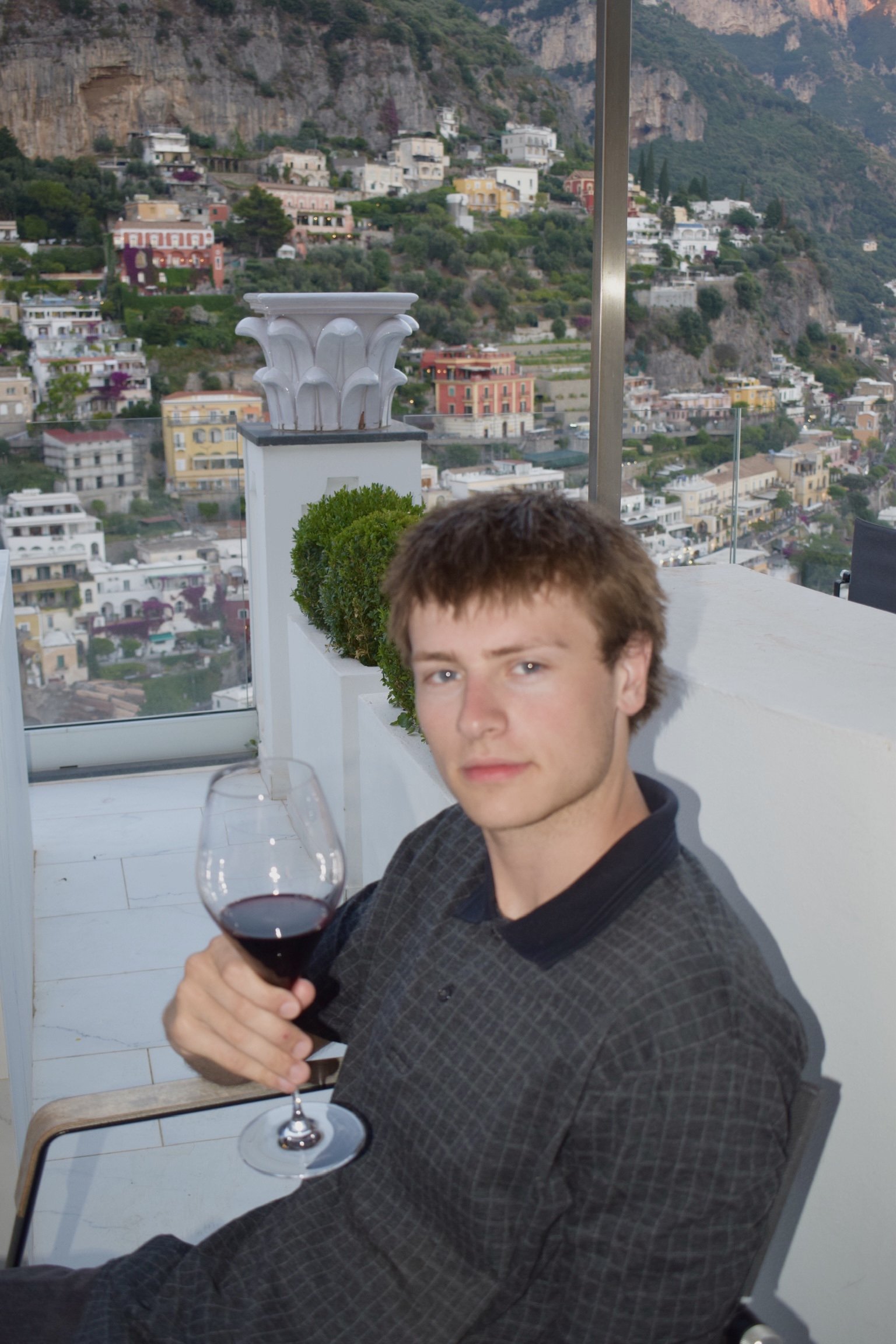 A young man with brown hair is sitting at an outdoor table, holding a glass of red wine, with a scenic hillside town in the background.