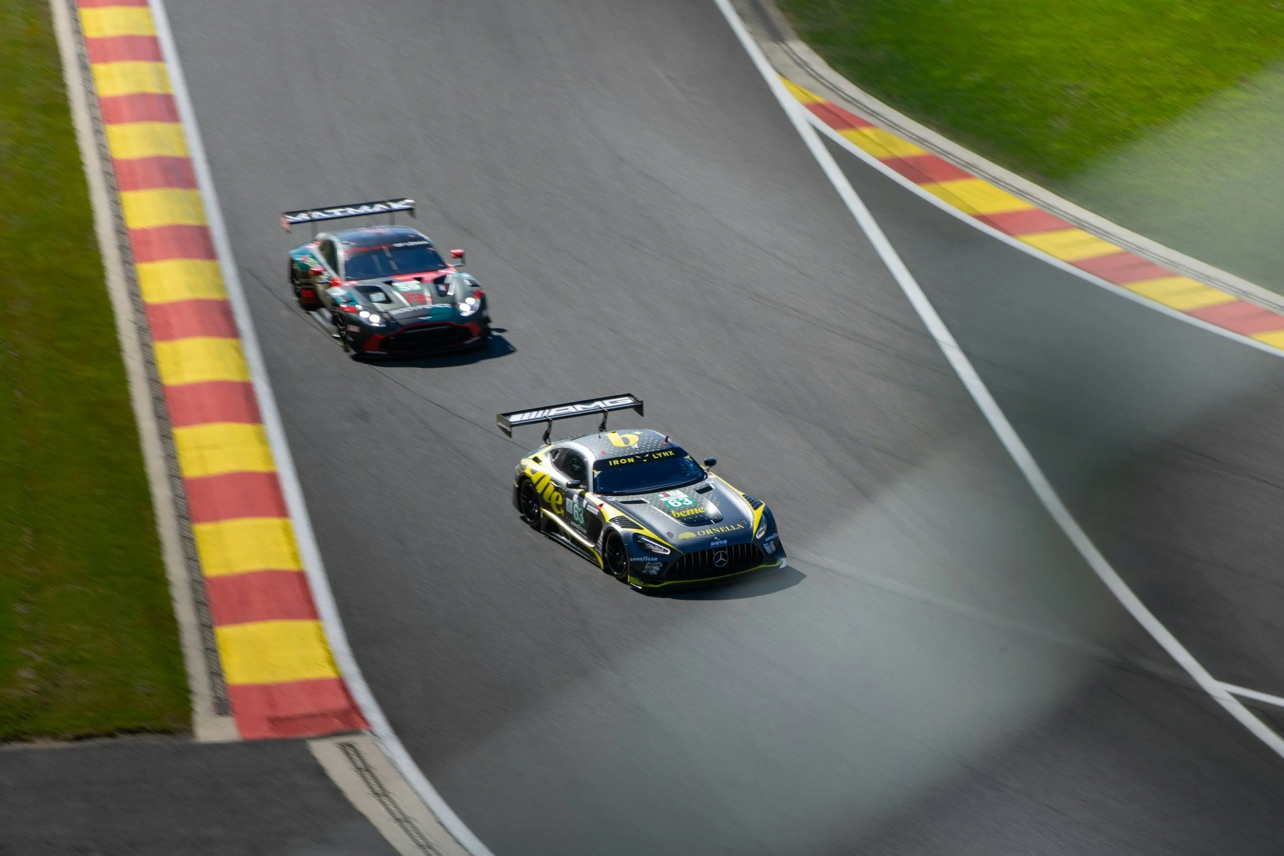 Une photo de voitures de course sur une piste de course en montée, entourée de bandes de peinture jaunes et rouges, avec de l'herbe verte sur les côtés.