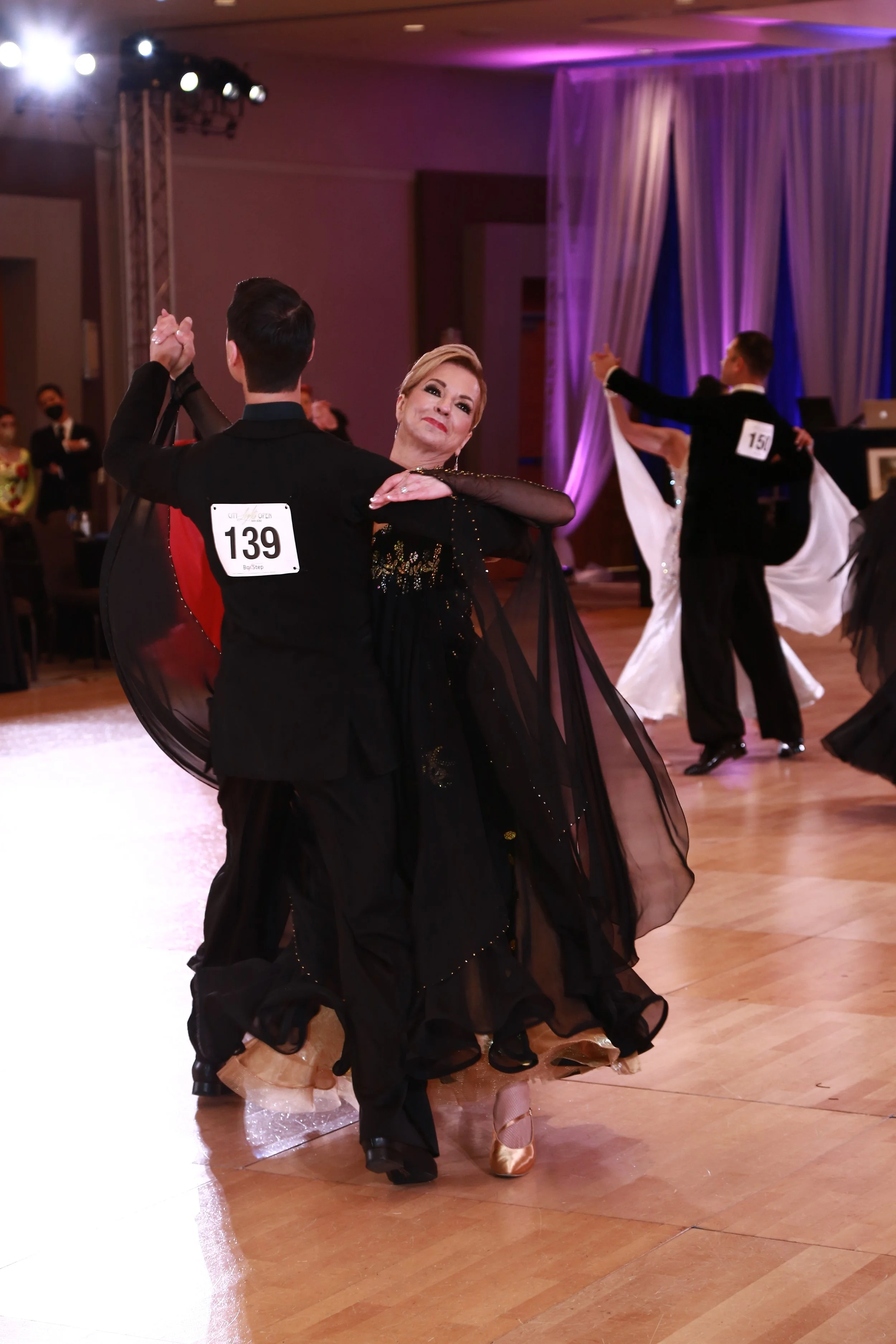 Couple dancing ballroom in elegant attire, with other couples in the background, under purple and pink lighting.