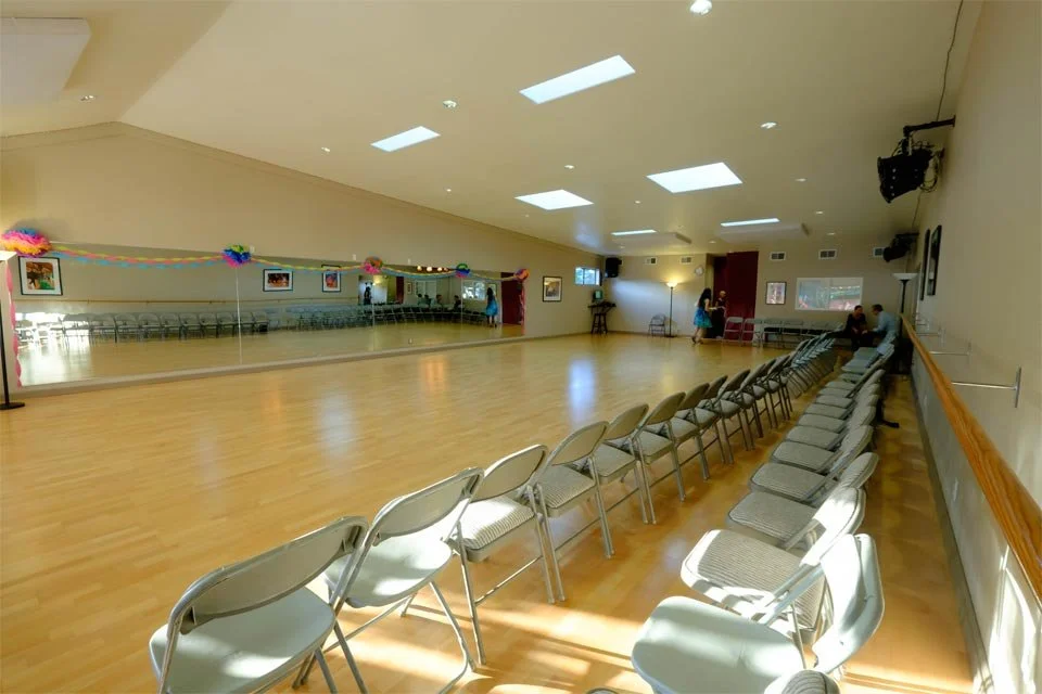 Empty dance studio with a row of chairs along the right side, some people at the back, and a mirrored wall decorated with colorful paper garlands.