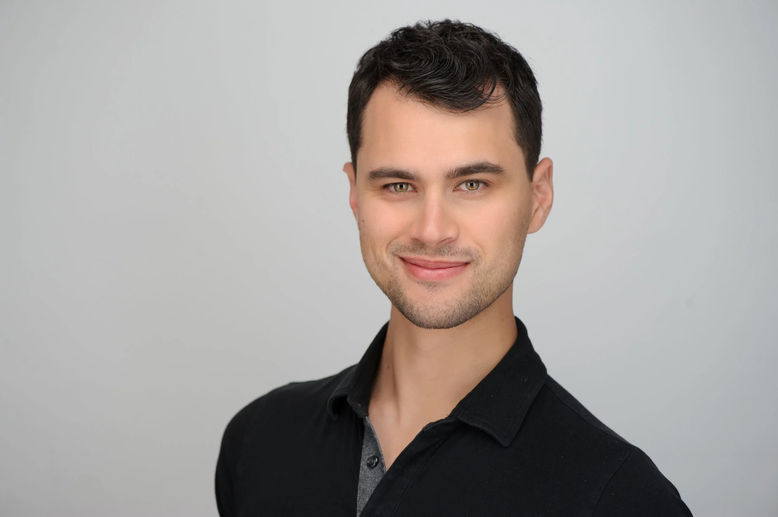 A young man with short dark hair, light complexion, and hazel eyes, smiling slightly, wearing a black shirt with a gray collar, against a plain light gray background.