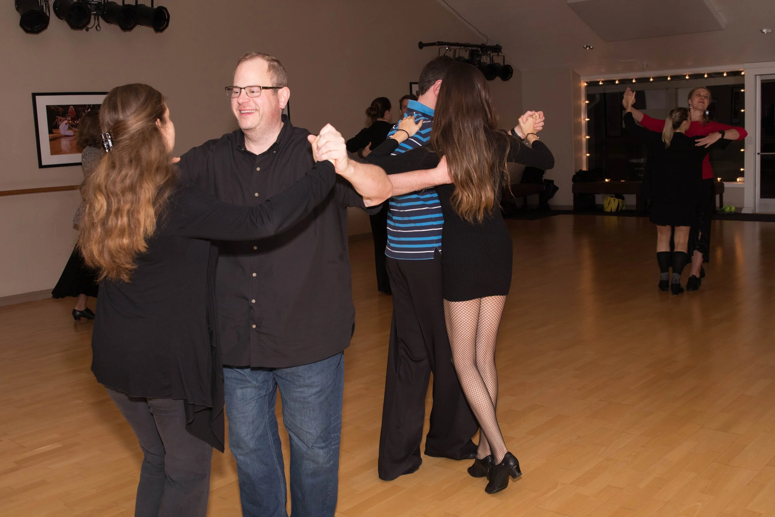 Couples dancing together in pairs in a dance studio, with two women in black dresses with fishnet stockings and heels, and men in casual attire, in a warmly lit room with wooden floors and a window at the back.
