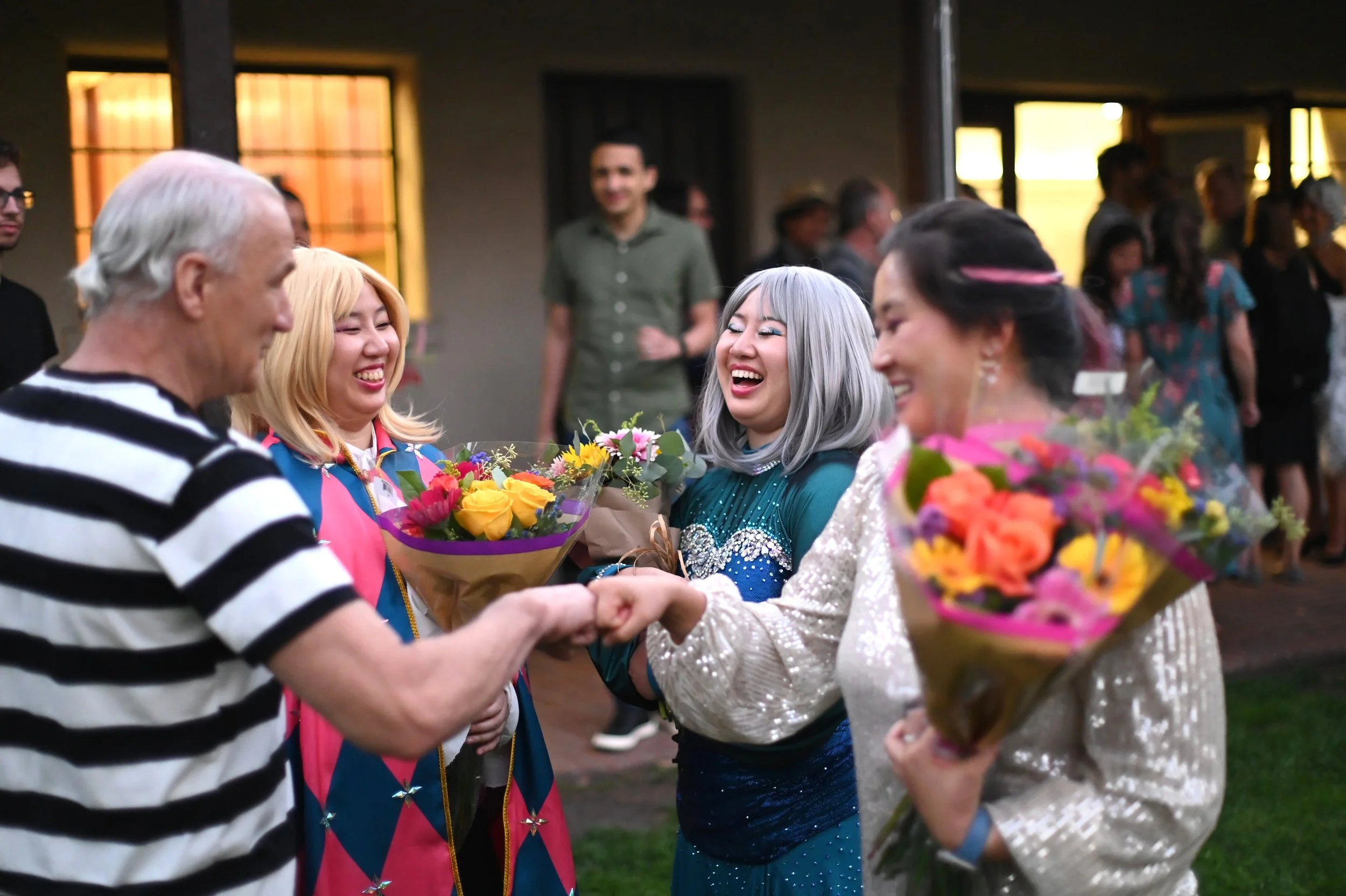 A group of people, including young women and an older man, exchanging flowers and shaking hands at a celebratory event in a well-lit indoor space. The women are dressed in colorful outfits and smiling.