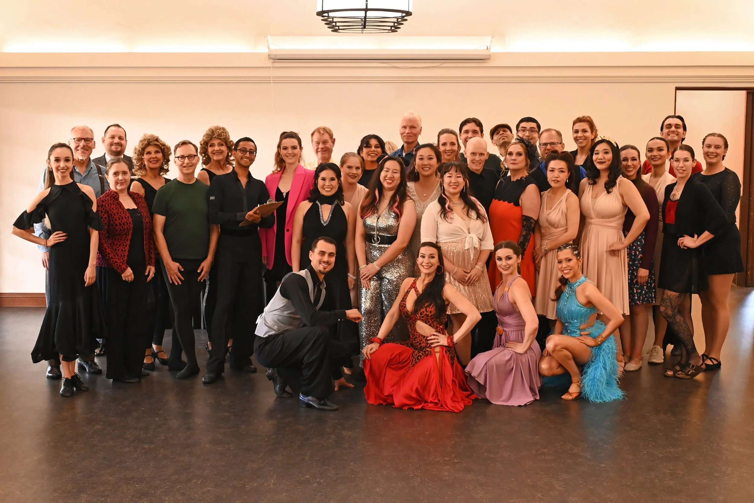 A large group of diverse people dressed in formal and colorful attire posing together indoors, some in dancing costumes, in front of a plain wall and ceiling lights.