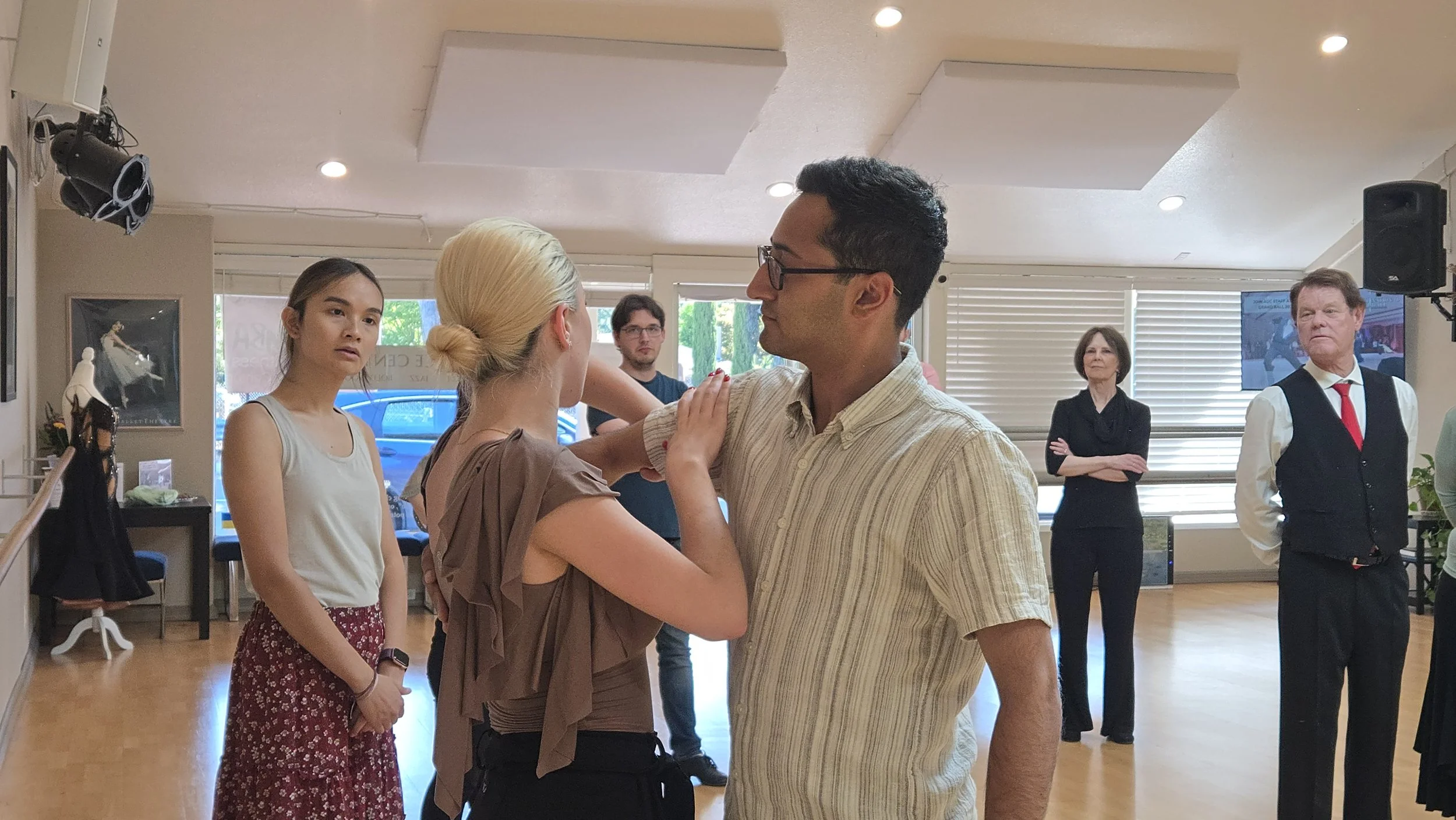 People dancing closely in a dance class or social event, with some watching in the background.