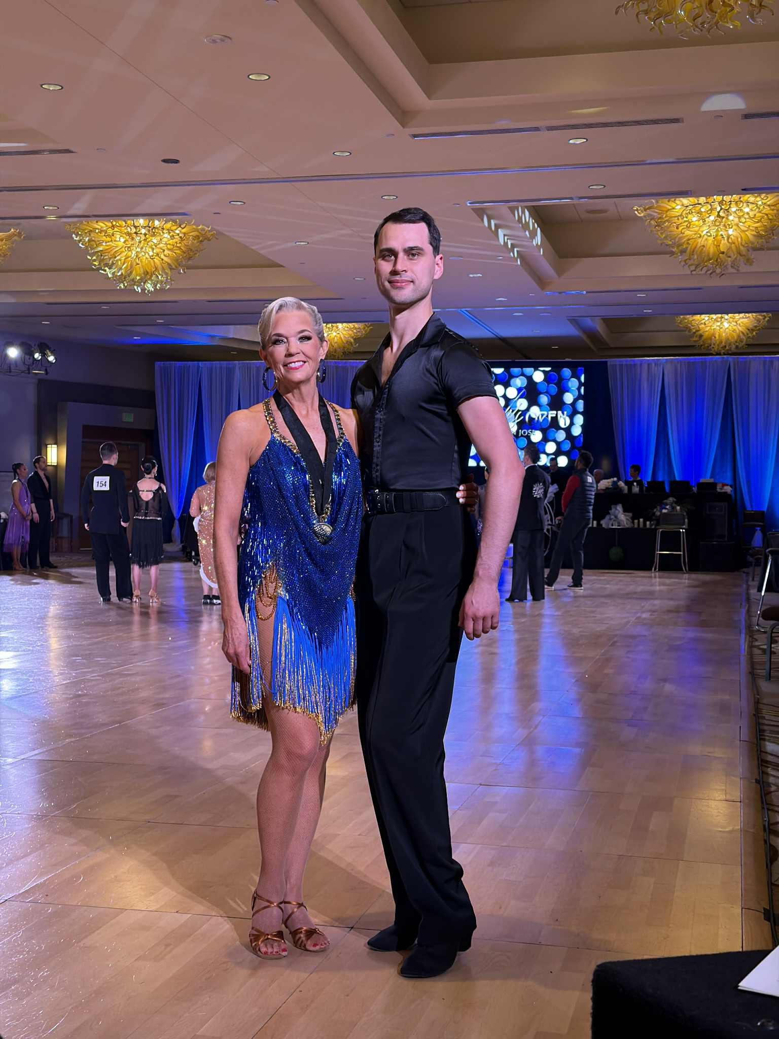A woman and a man in dance attire pose together on a ballroom dance floor, with other dancers and event staff in the background.