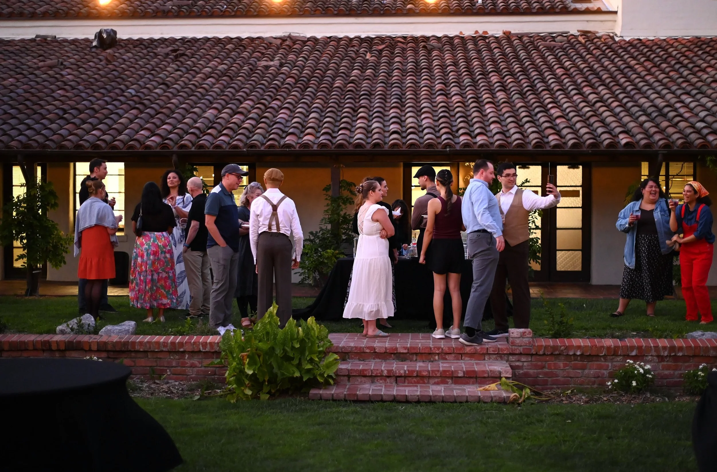 A group of people gathered outdoors on a lawn, socializing and taking photos in front of a yellow house with a tiled roof and lit windows at dusk.