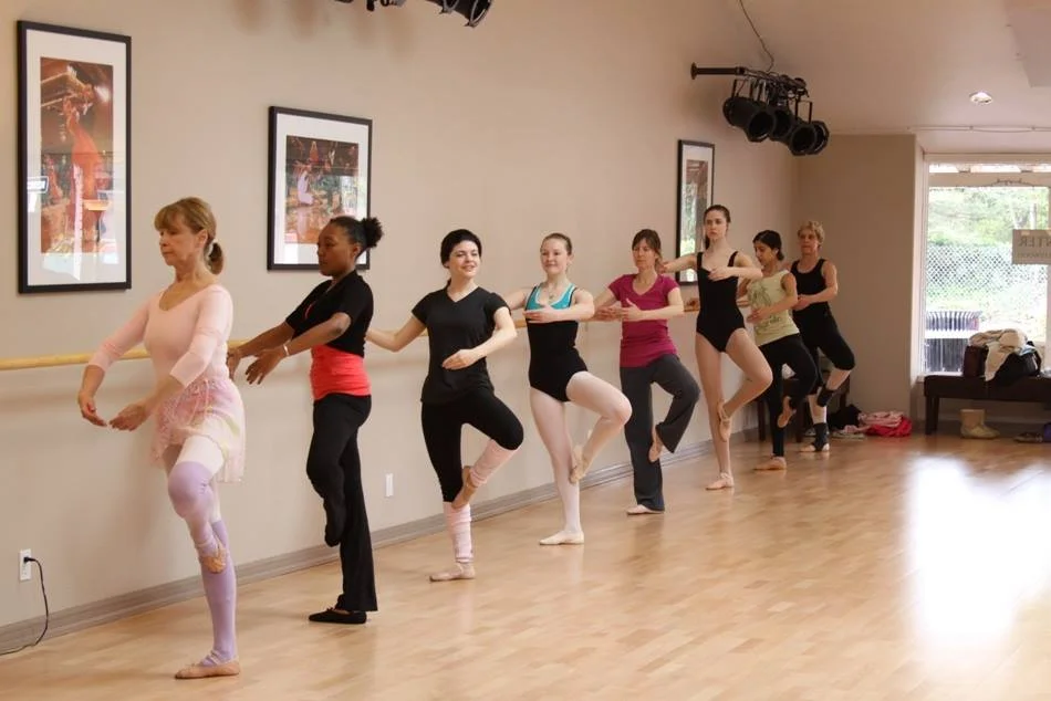 Ballet class with seven students holding onto a ballet barre in a dance studio, practicing ballet poses.