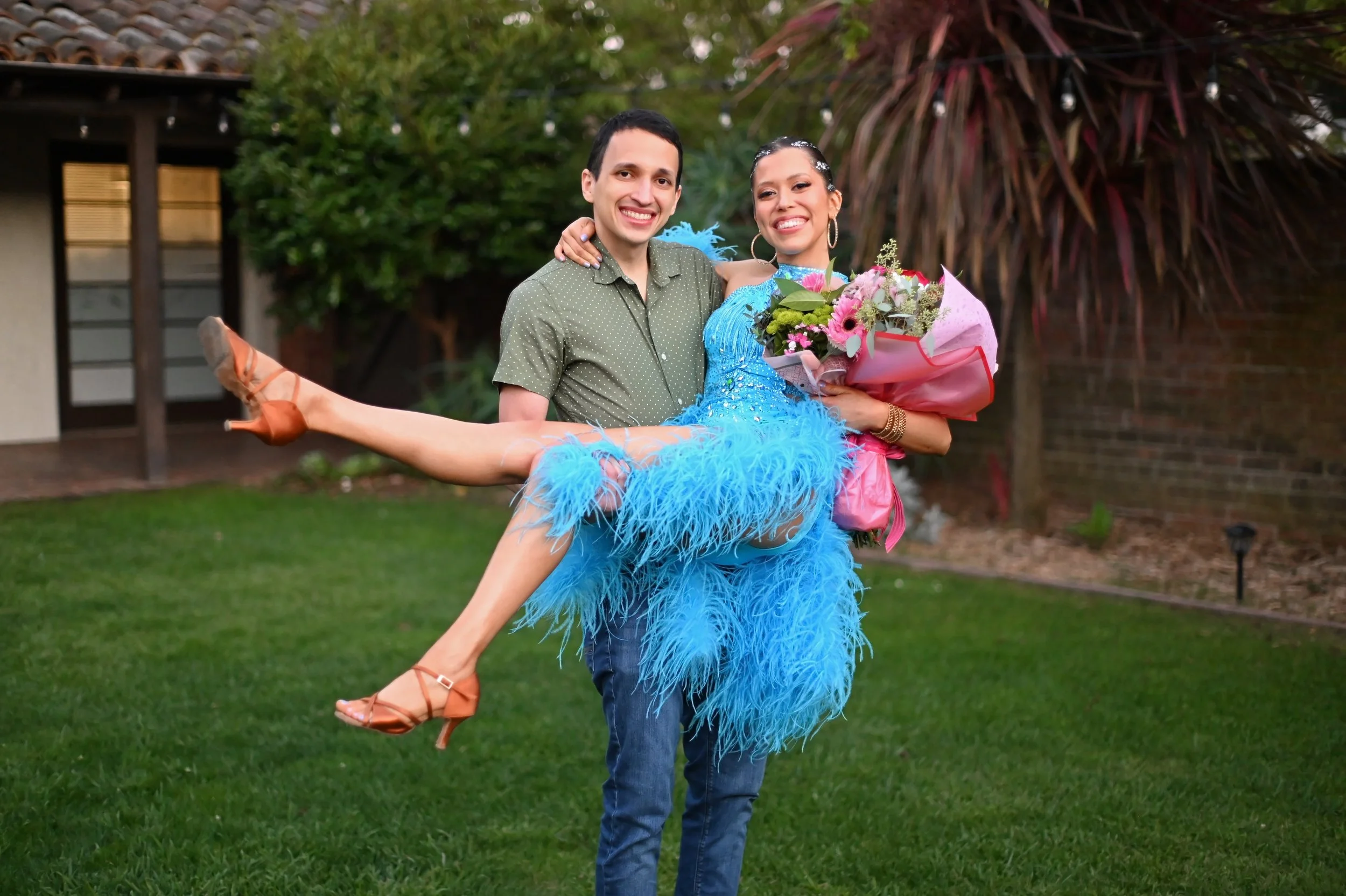 A man in a green shirt lifting a woman in a bright blue dress with feather decorations, holding a bouquet of flowers, outdoors in a backyard with trees and a building in the background.