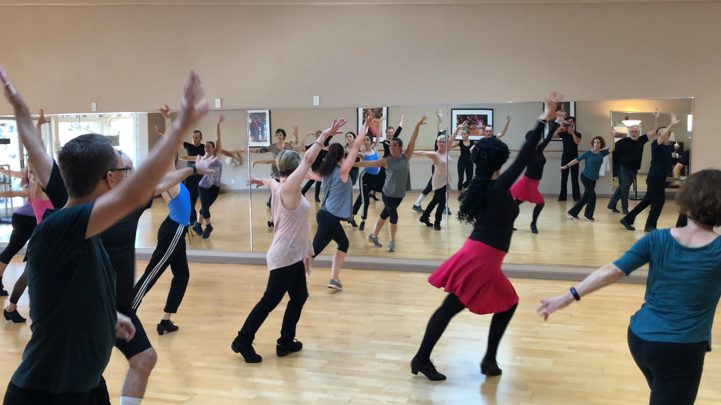 A diverse group of people participating in a dance class in a studio with wooden floors and large mirrors, raised arms, moving rhythmically.