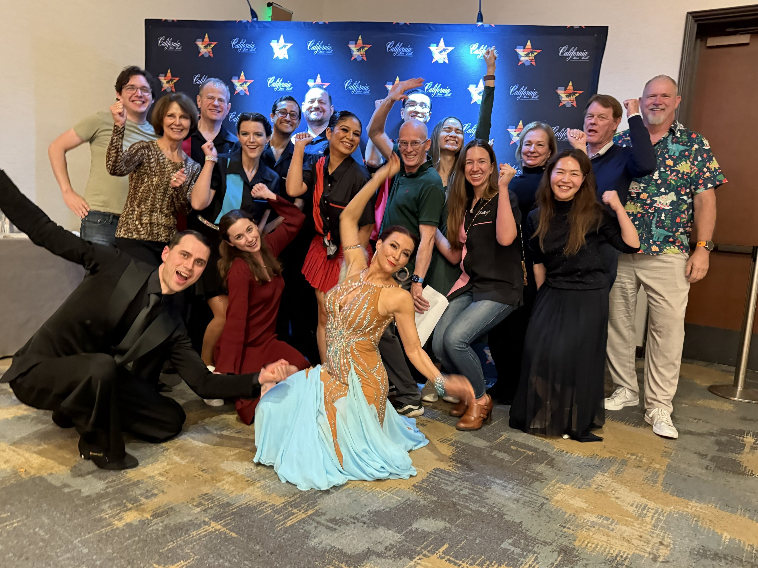 Group of people celebrating and smiling, with some raising fists or fists in the air, standing in front of a backdrop with stars and the word 'California'. One person at the front is dressed in a colorful dress and posed in a dance position.