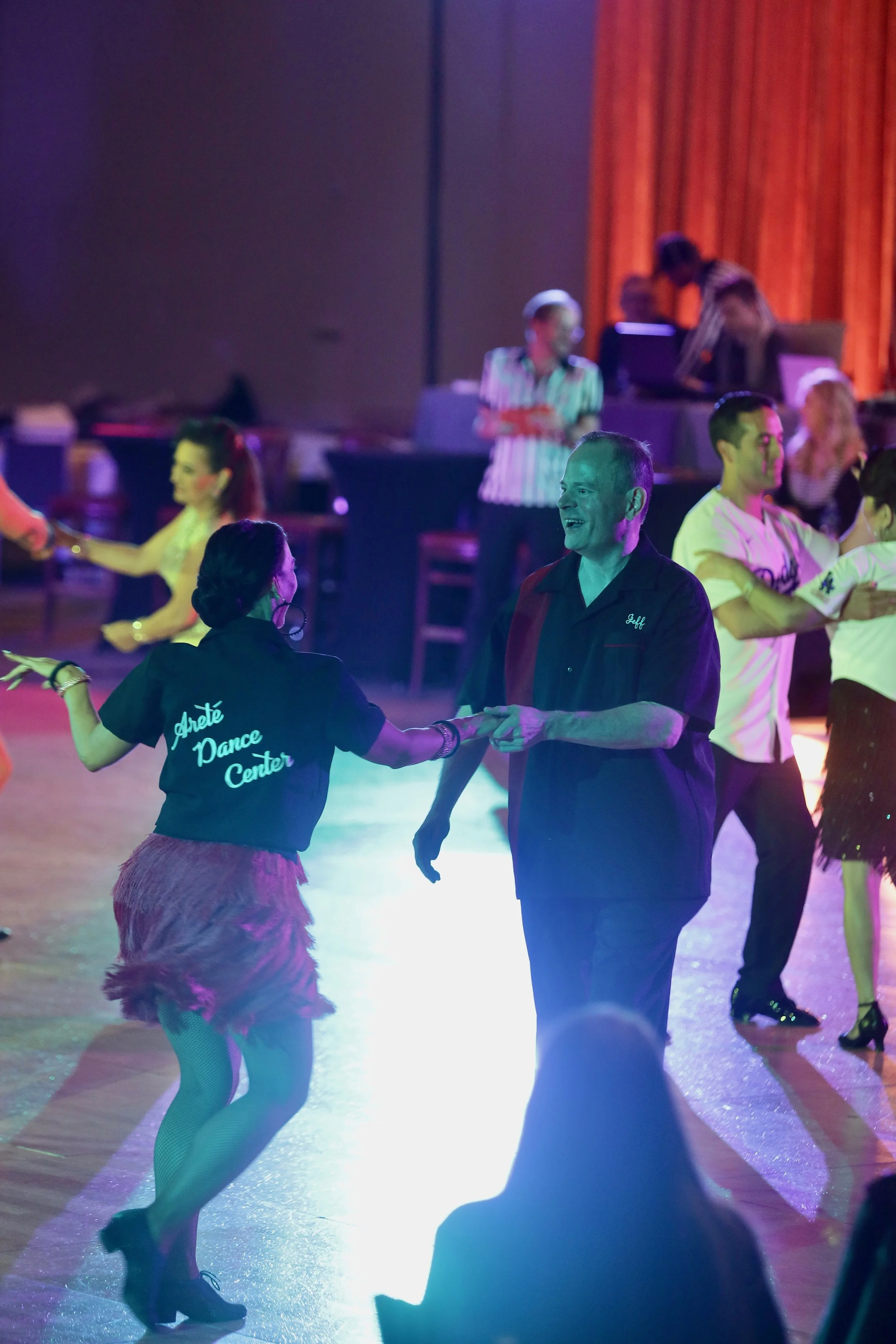 Couples dancing on a brightly lit dance floor with colorful lights, in a lively social setting.