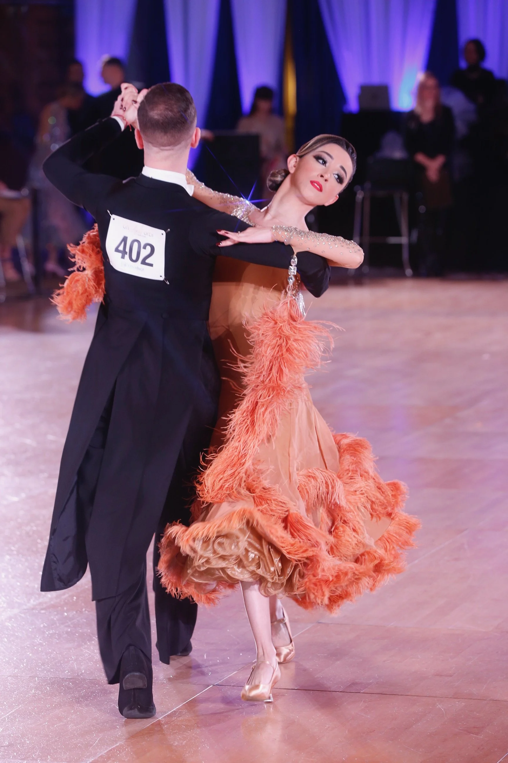 A couple performing a ballroom dance, with the woman wearing an elegant orange dress with feathers and the man dressed in a black suit with a number 402 on his back.