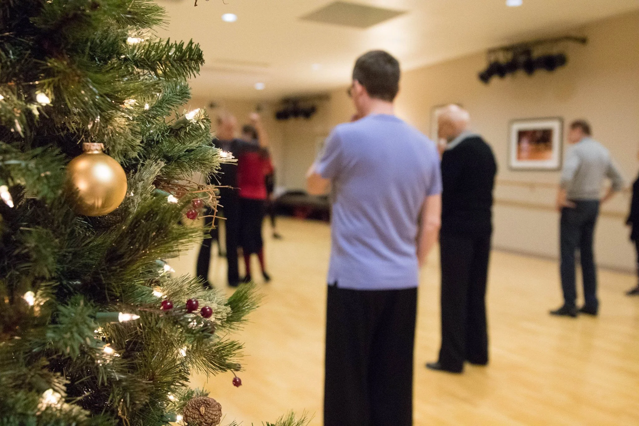 A decorated Christmas tree with lights, gold ornaments, red berries, and pinecones on the left side of the image, with a group of people gathered in a softly lit room in the background, some engaged in conversation.