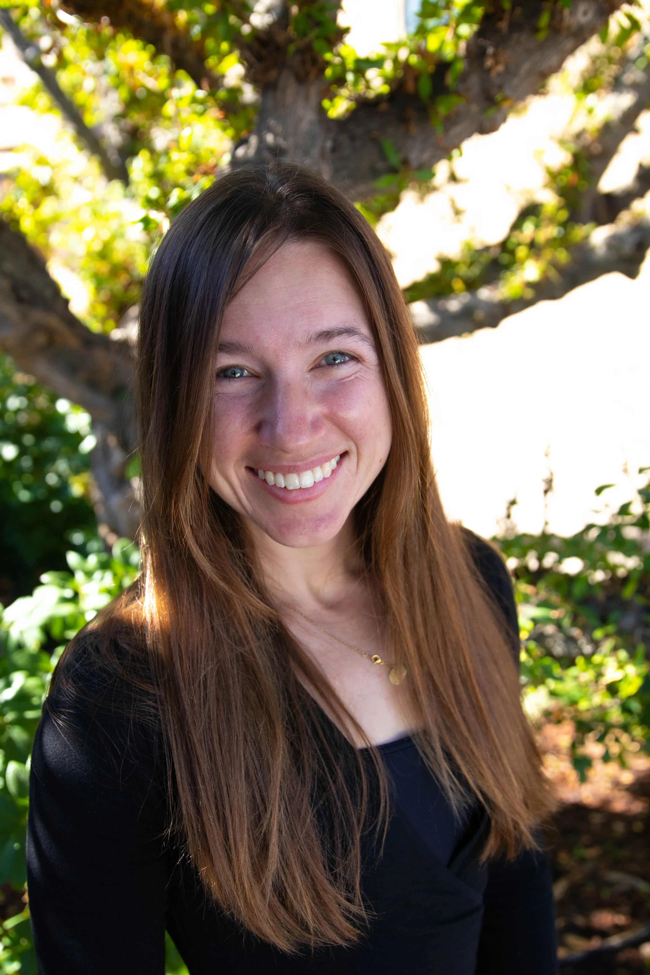 A smiling woman with long brown hair wearing a black top and a gold necklace, standing outdoors with a tree and green foliage in the background.