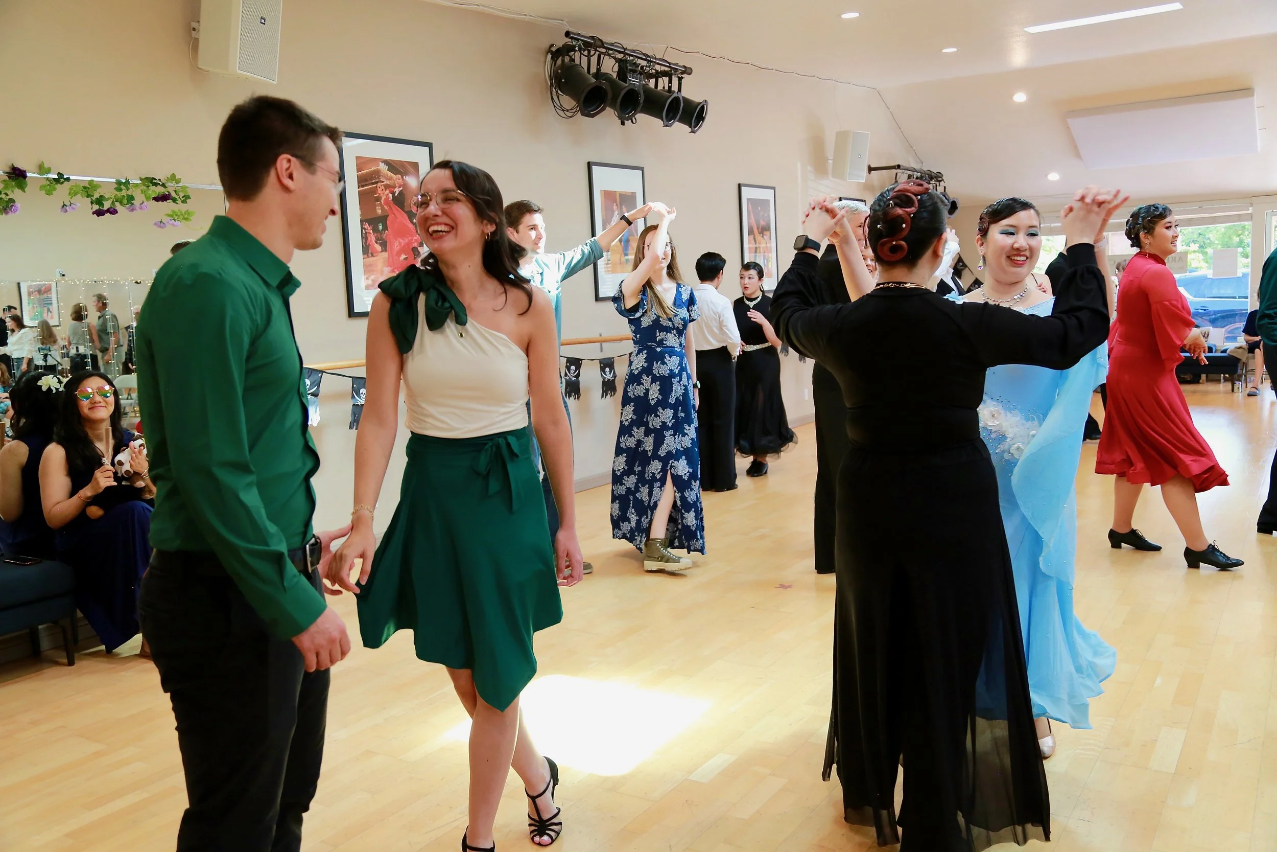 People dancing in a spacious room with framed pictures on the wall, some sitting and watching, others dancing in colorful dresses and suits.