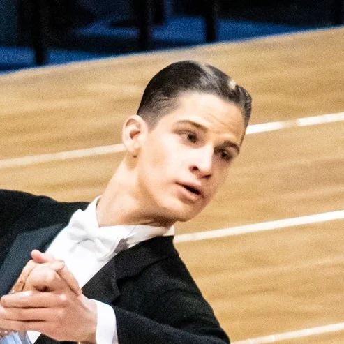 A young man in a tuxedo posing with his hands clasped together in front of him, standing on a wooden floor.