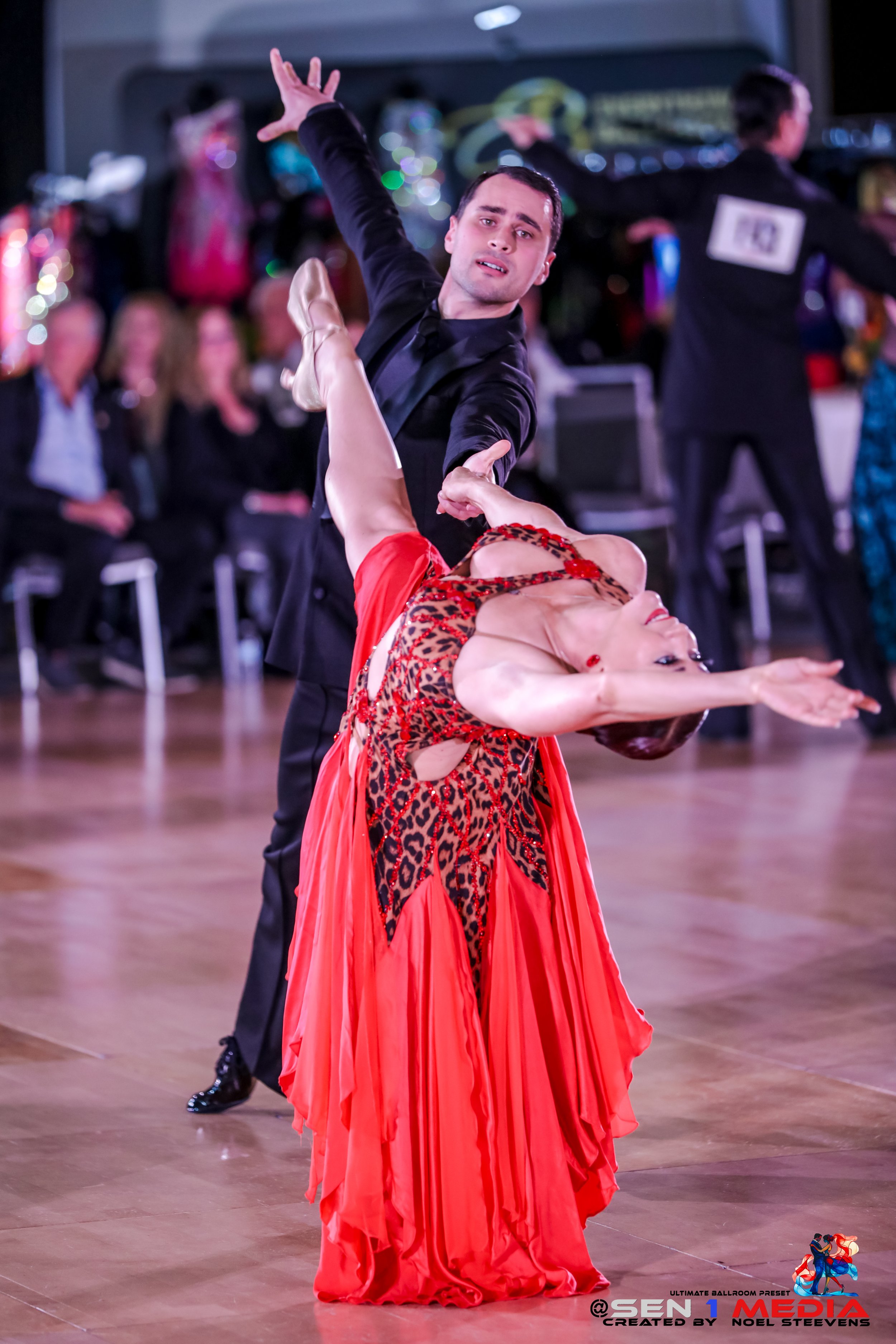 A male ballroom dancer in a black suit and a female ballroom dancer in a long, flowing red dress with leopard print details performing a dance routine on a wooden floor with an audience in the background.