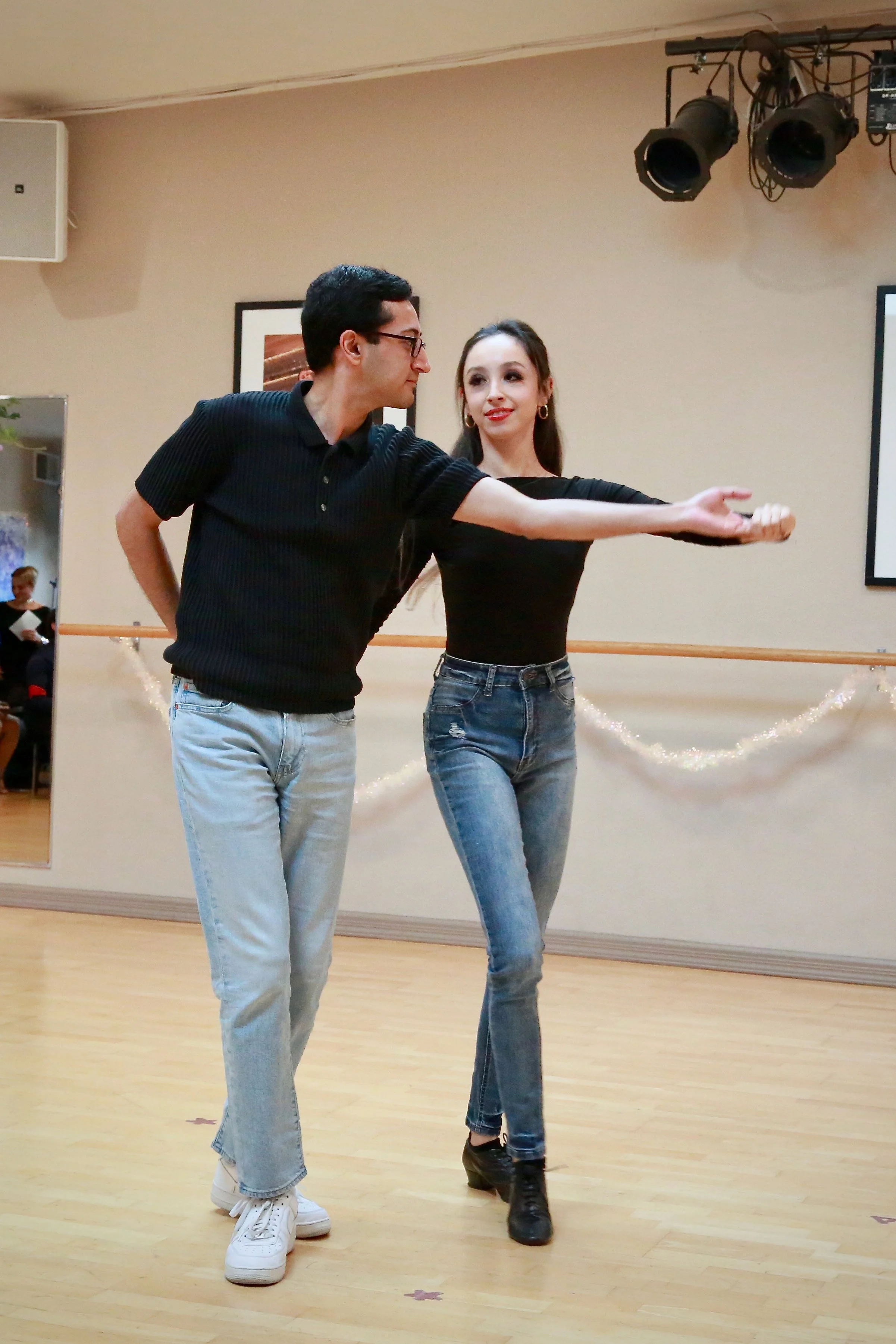 A man and woman dancing together in a dance studio, with a mirror on the wall and professional stage lights overhead.