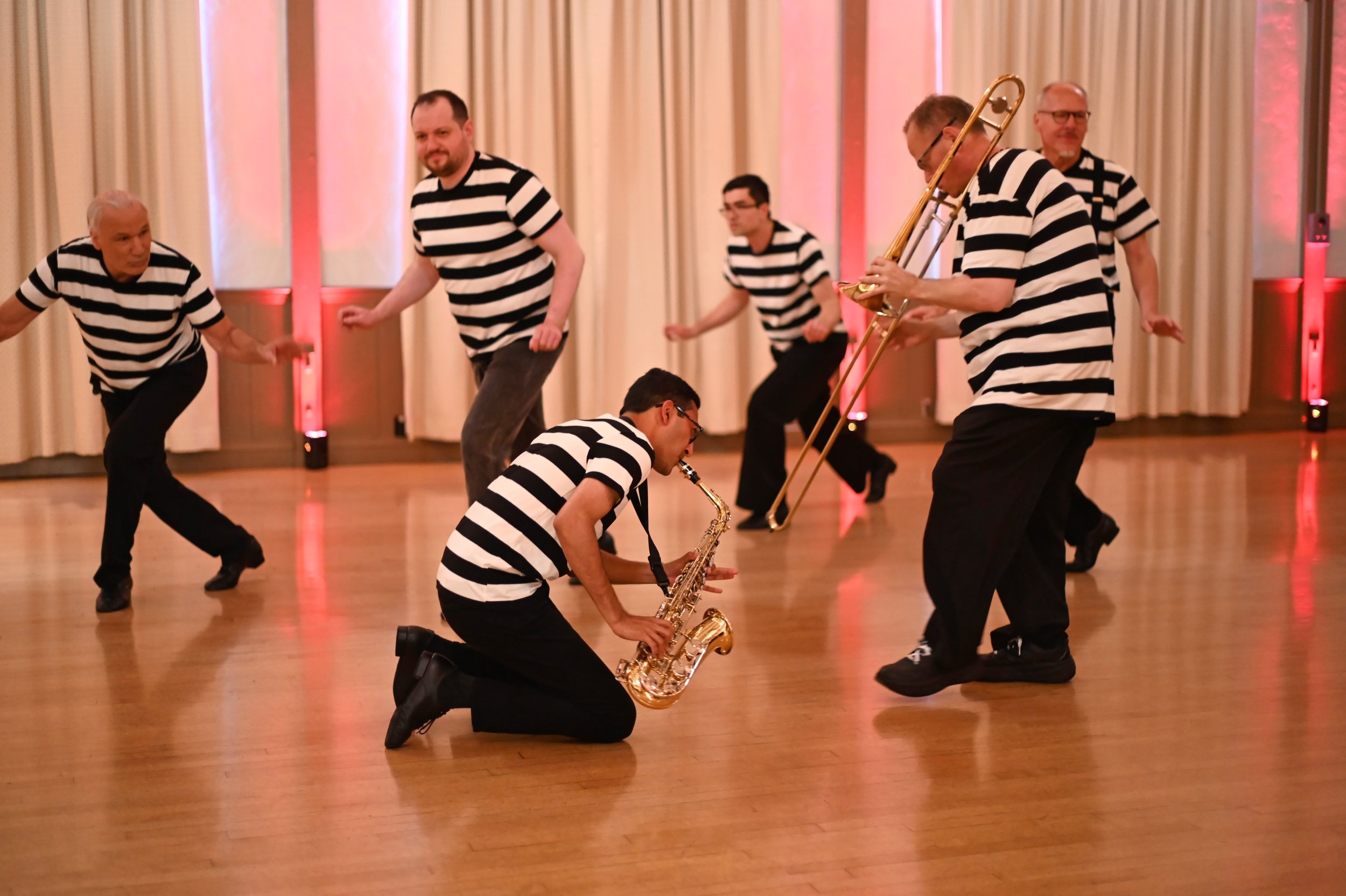 Men in black and white striped shirts dancing and playing musical instruments in a decorated hall.