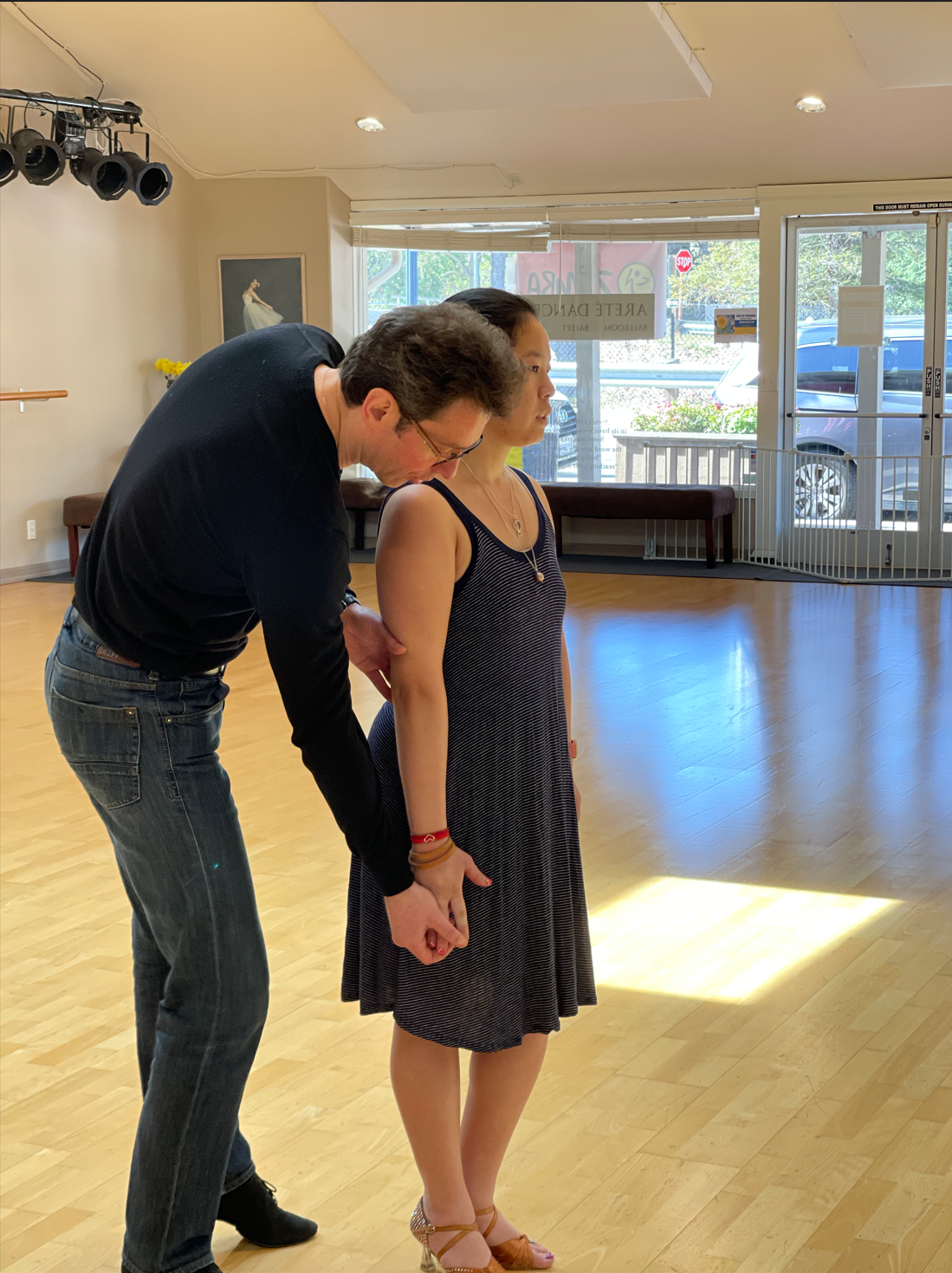 A man is assisting a woman with a dance move in a dance studio. The man leans forward, holding her hand, while she stands facing forward. The room has large windows, wooden floors, and minimal decor.