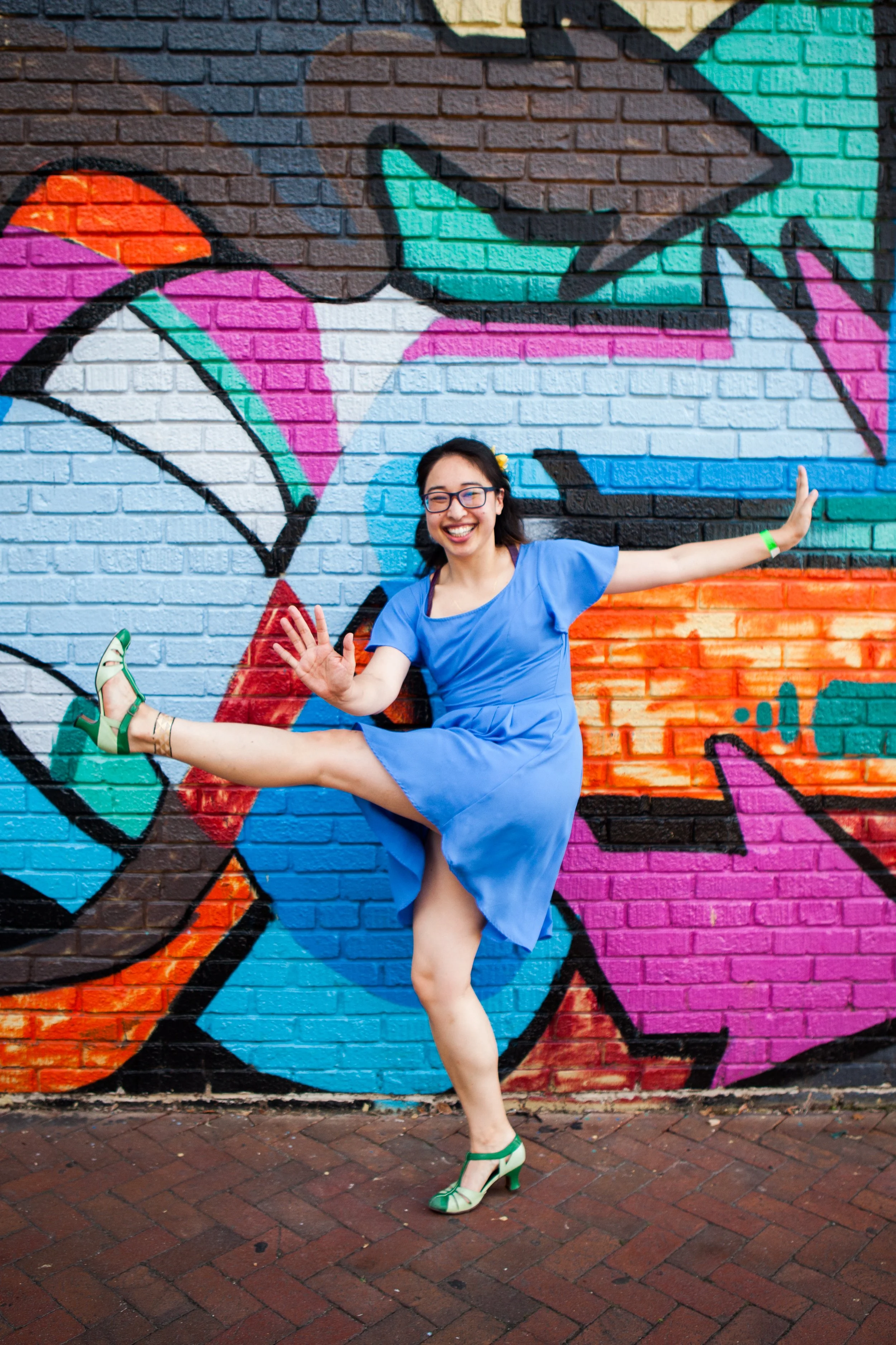 A woman in a blue dress and green high heels is performing a dance pose in front of a colorful graffiti wall, smiling.