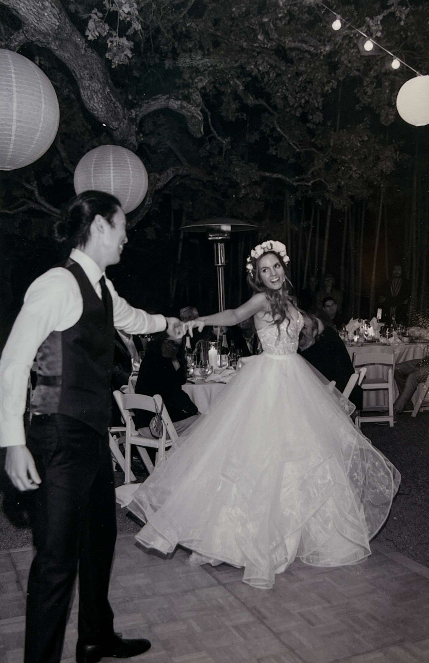 A couple dancing at a wedding reception outdoors at night, with the bride wearing a white wedding gown with a full skirt and a floral crown, and the groom in formal attire. Lanterns hang from trees above, and seated guests and tables are visible in the background.