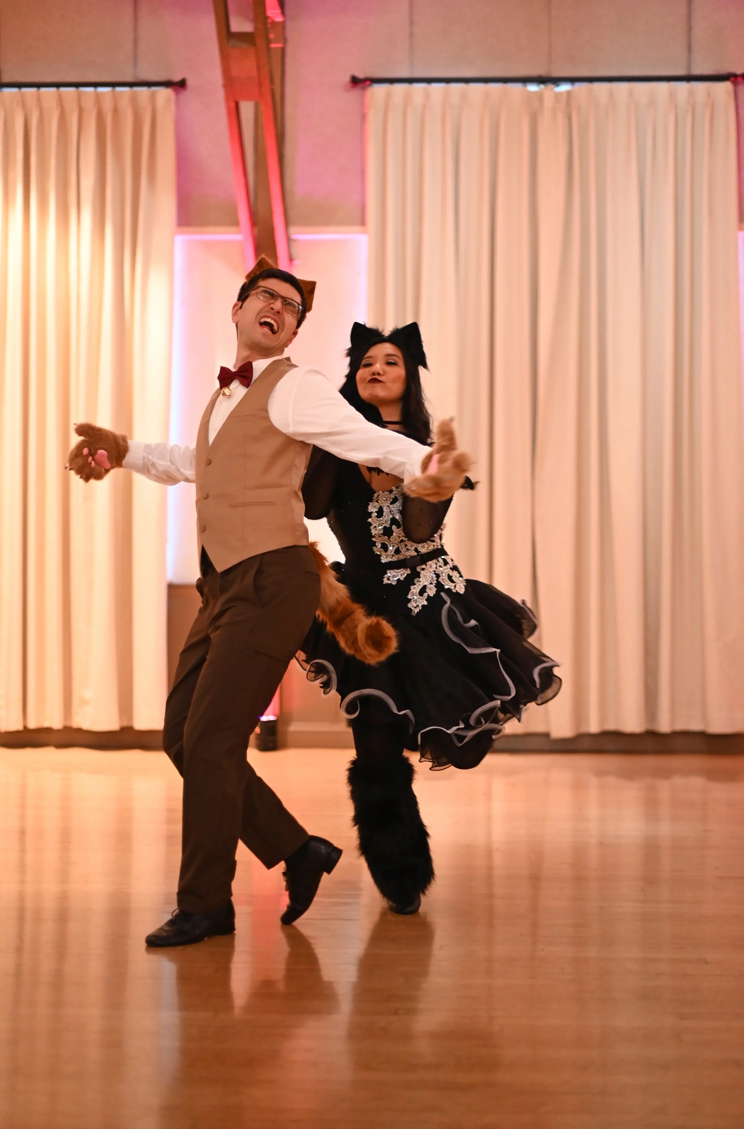 Two people dancing in costumes that resemble cats, in a room with beige curtains and wooden stage flooring.