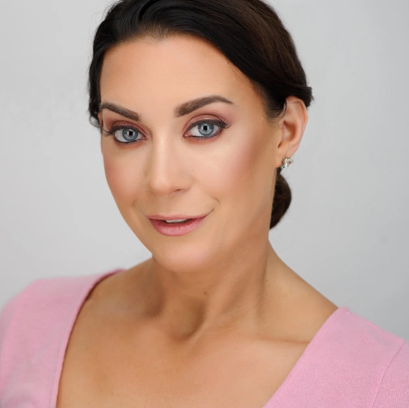 A woman with dark hair styled in an elegant updo, wearing light makeup and a pink top, smiling softly against a plain background.
