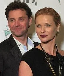 A man and a woman standing close together, posing for a photo at an event. The man has curly dark hair and is wearing a dark suit with a white shirt. The woman has blonde hair styled back, wearing a black top and a necklace.