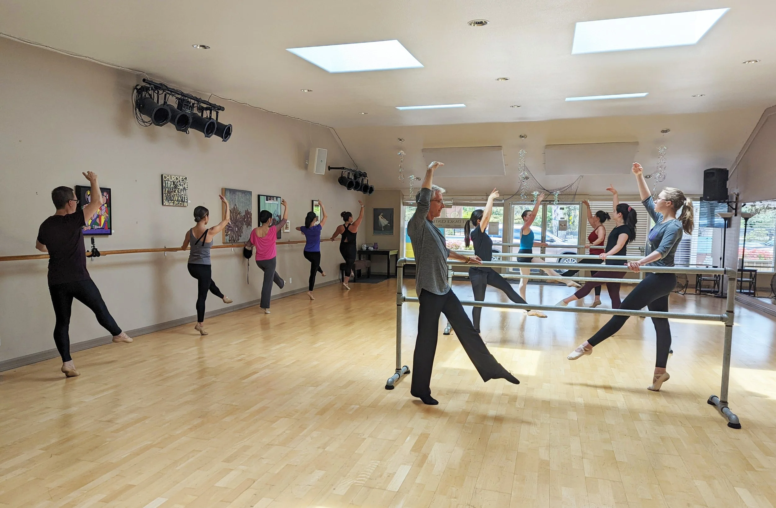 A group of ballet dancers practicing in a dance studio with mirrors, wooden floors, and dance barre. The dancers are standing on one leg with the other leg extended behind them.