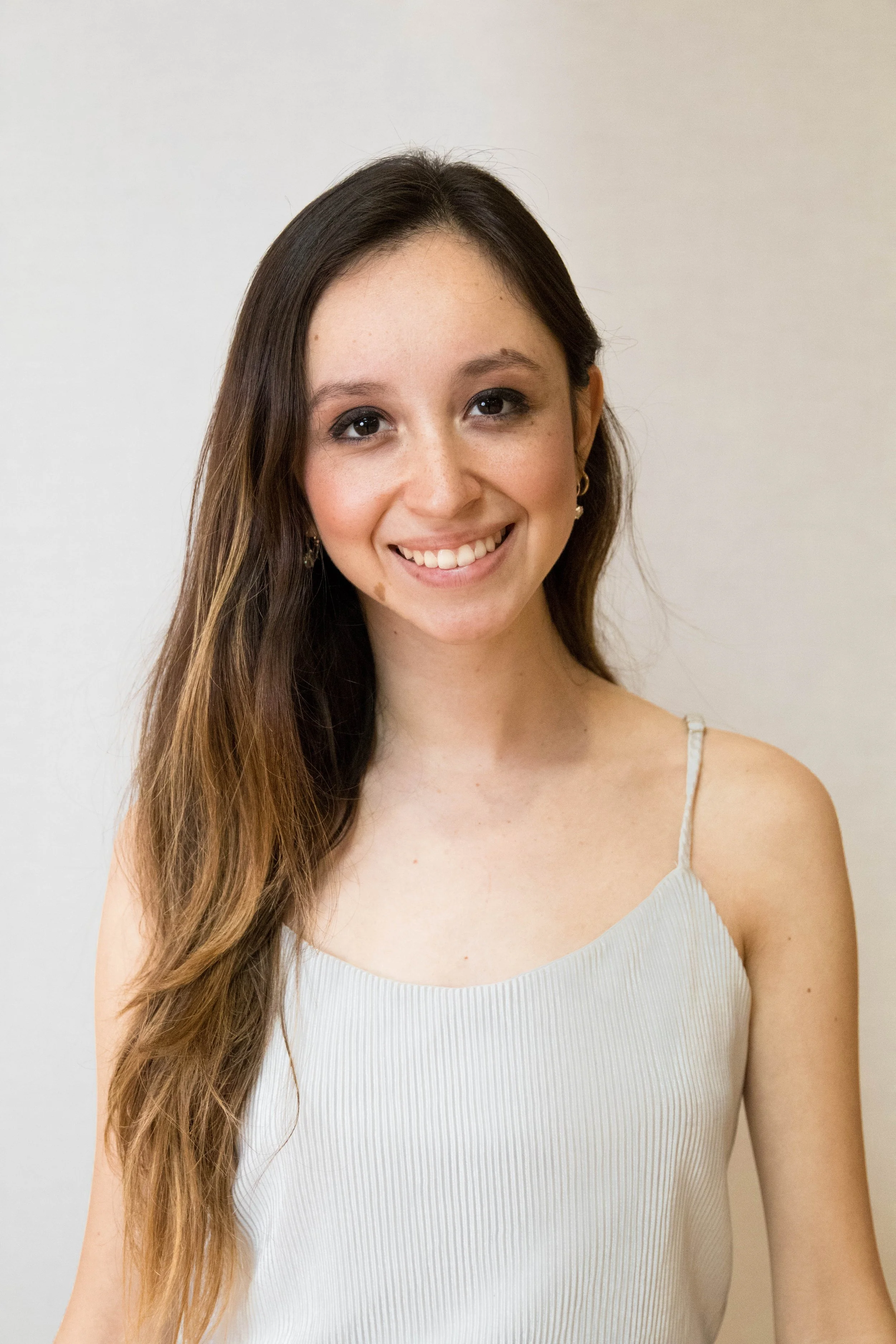 A young woman with long brown hair, wearing a light-colored, sleeveless top, smiling, standing against a plain, off-white background.