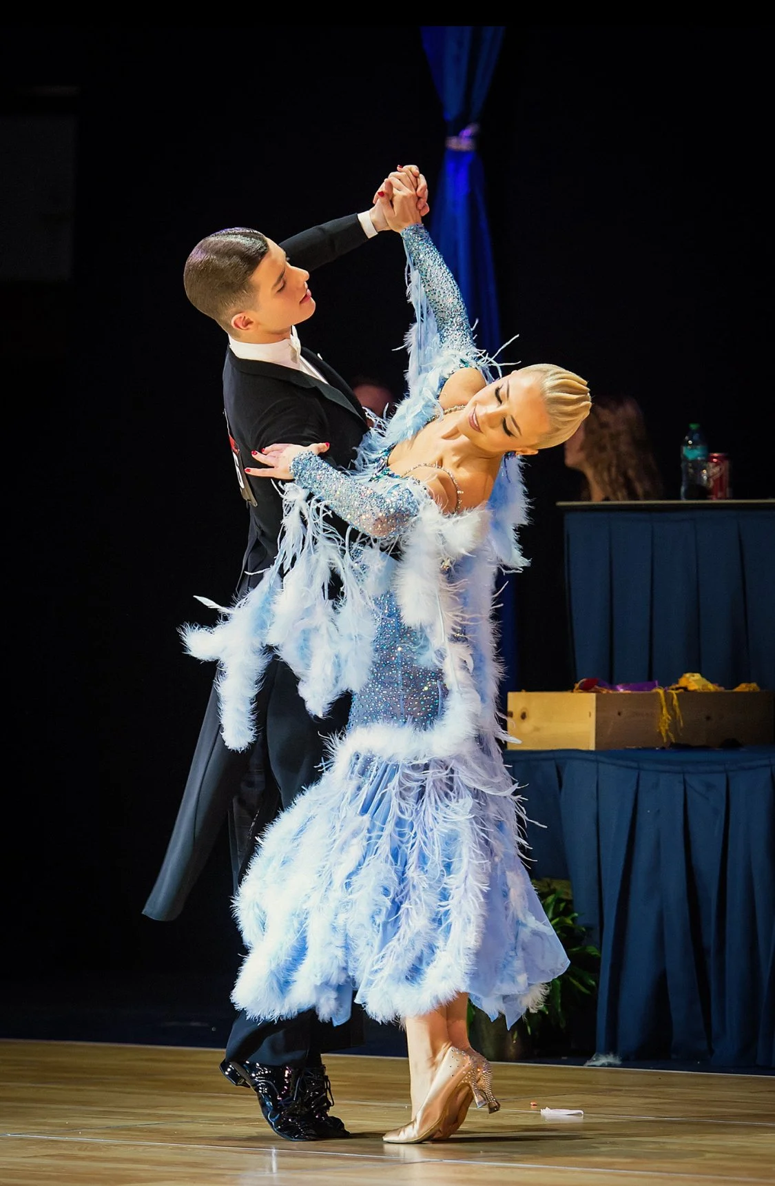 A ballroom dance couple in formal attire performing on stage. The woman wears a sparkly blue dress with feathers, and the man wears a black suit.