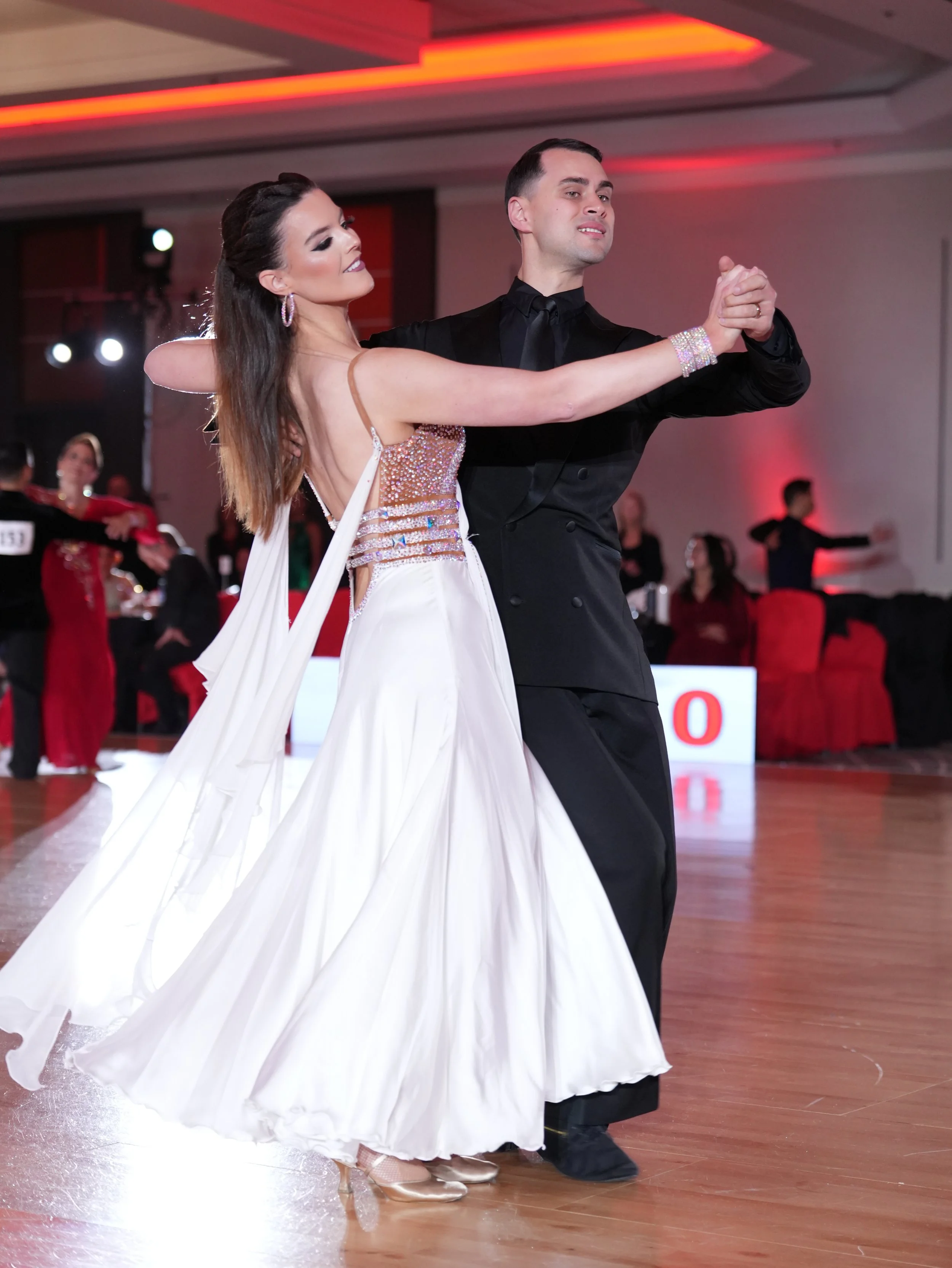 Couple dancing ballroom dance in formal attire, woman in a white gown with sequins, man in a black tuxedo, with other dancers in the background.