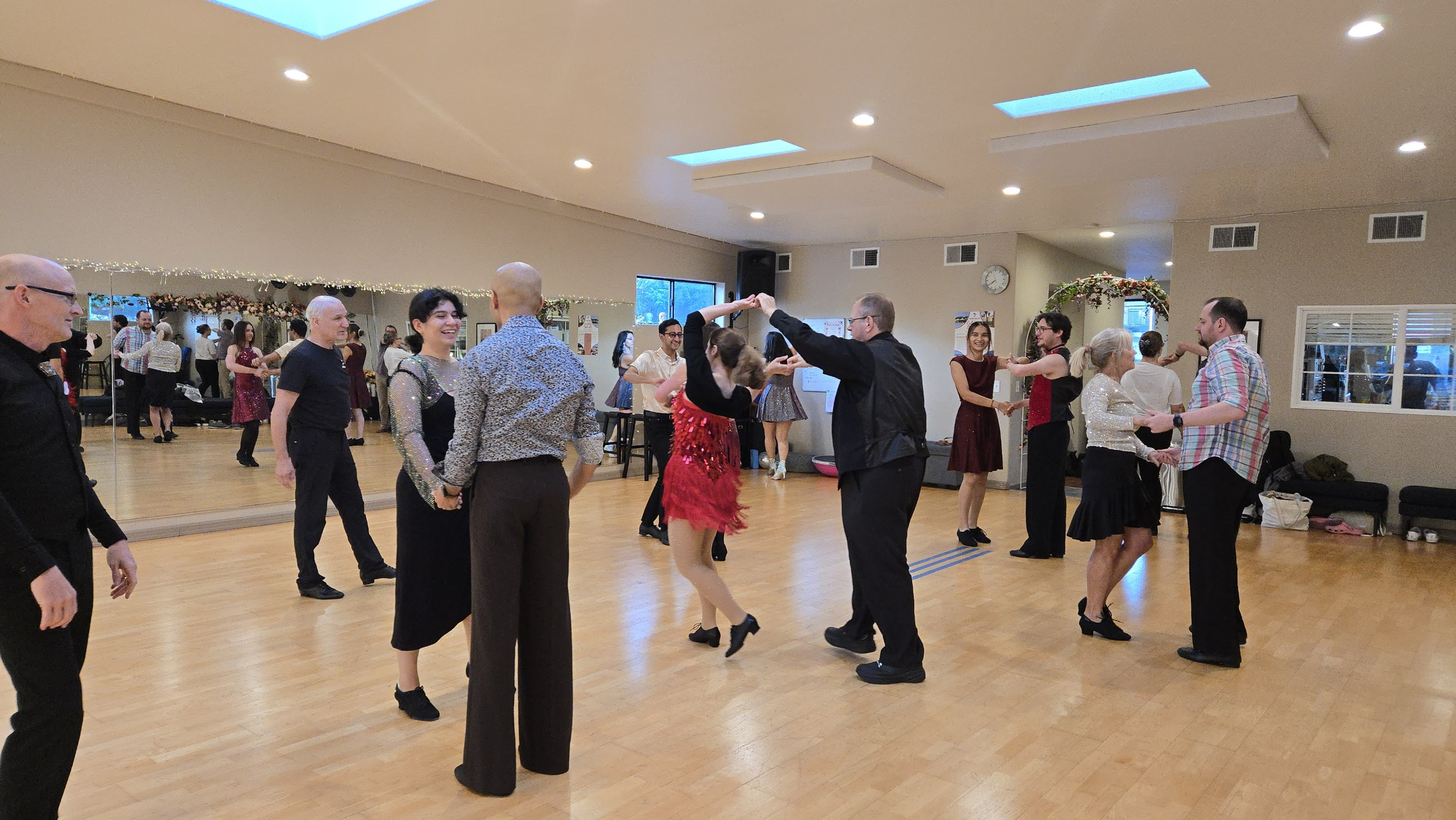 People dancing in pairs in a dance studio, with a mirror on one wall and decorated with hanging lights and floral decorations.