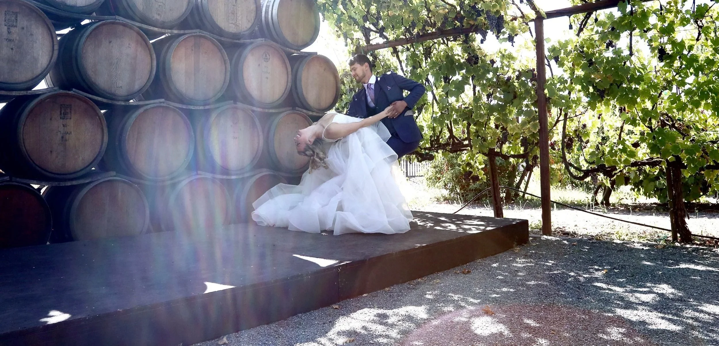 A groom and bride in wedding attire dancing outside among grapevines and stacked wine barrels.