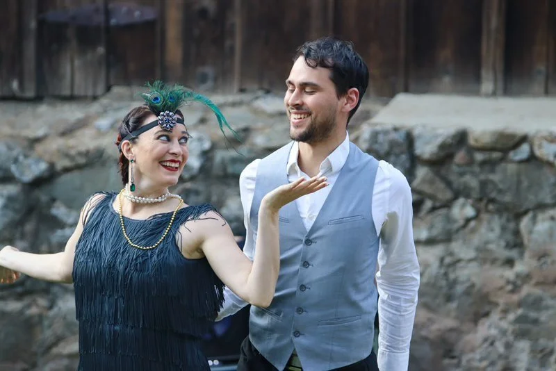 A woman in a 1920s flapper dress and a man in a vest dancing together outdoors with a rocky background.